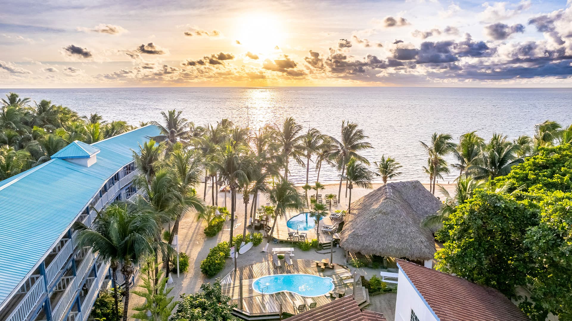 Aerial view of a tropical beach resort at sunset, featuring palm trees, pools, and the ocean.