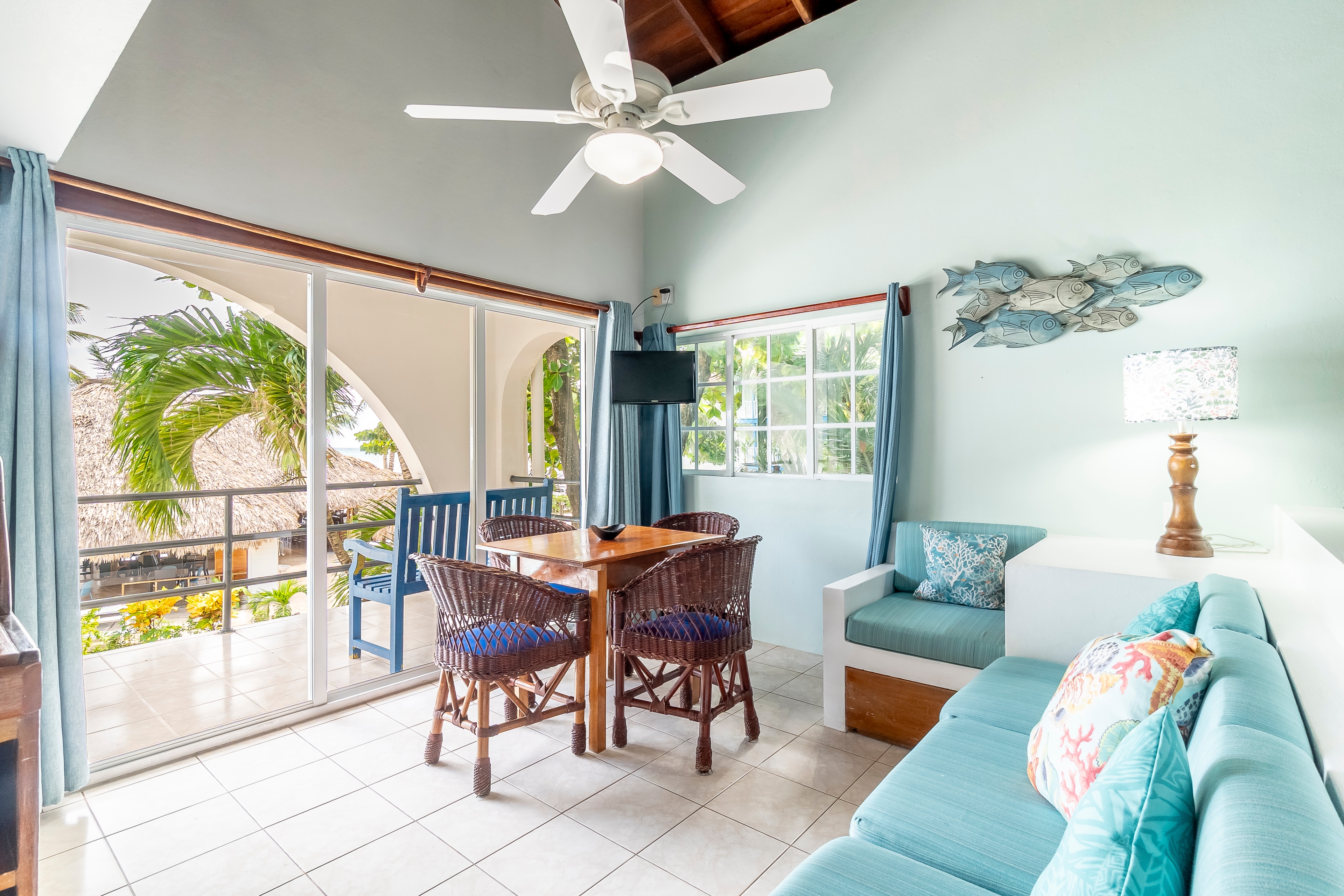 Brightly lit tropical suite living area featuring a light blue sofa, dining table, vaulted ceiling with a fan, and sliding glass doors opening to a balcony.