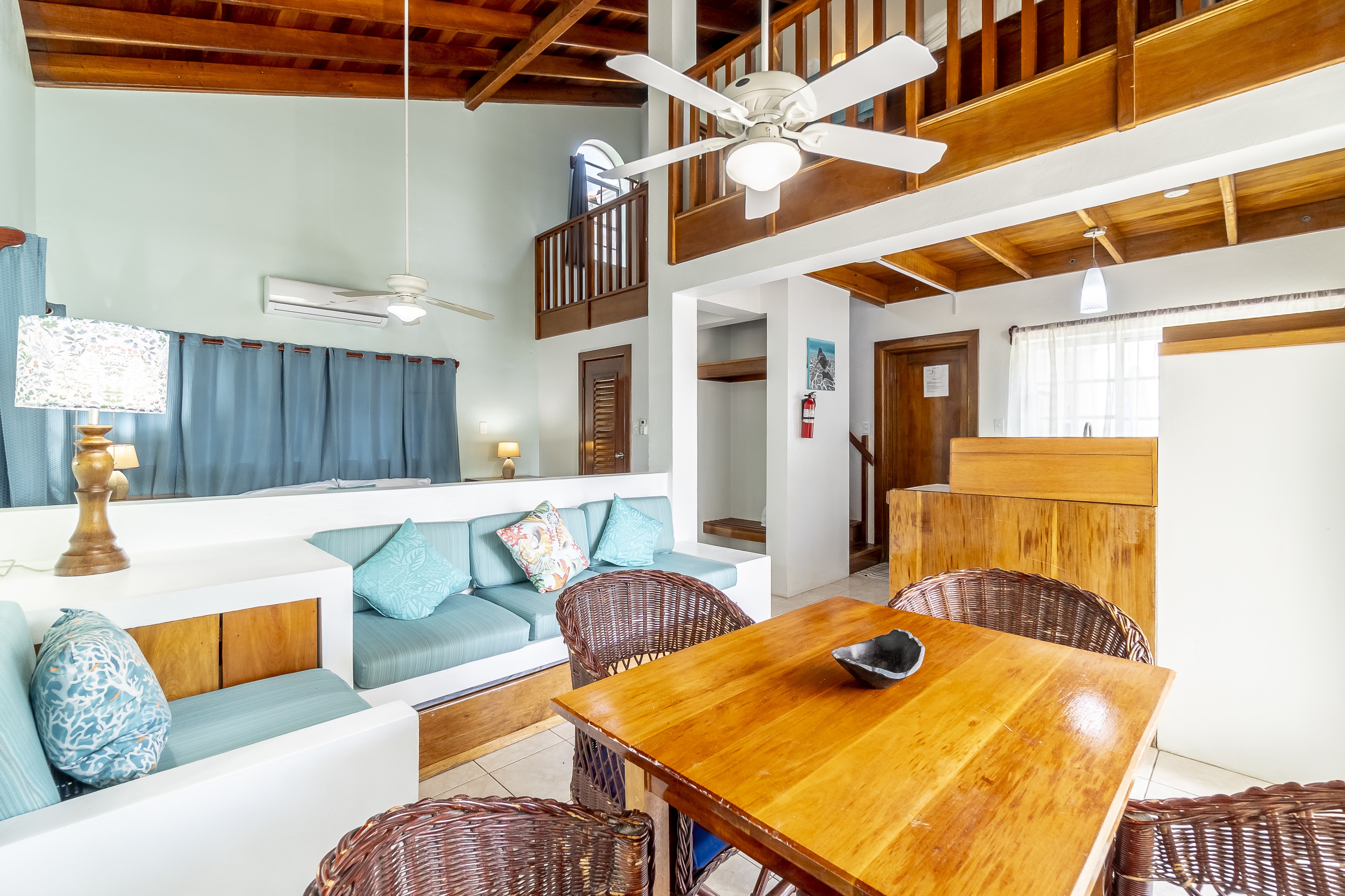 Spacious living area of a tropical suite featuring a vaulted wood-beam ceiling, a wooden dining set, and light blue sofas, with a view of an upper-level sleeping loft.