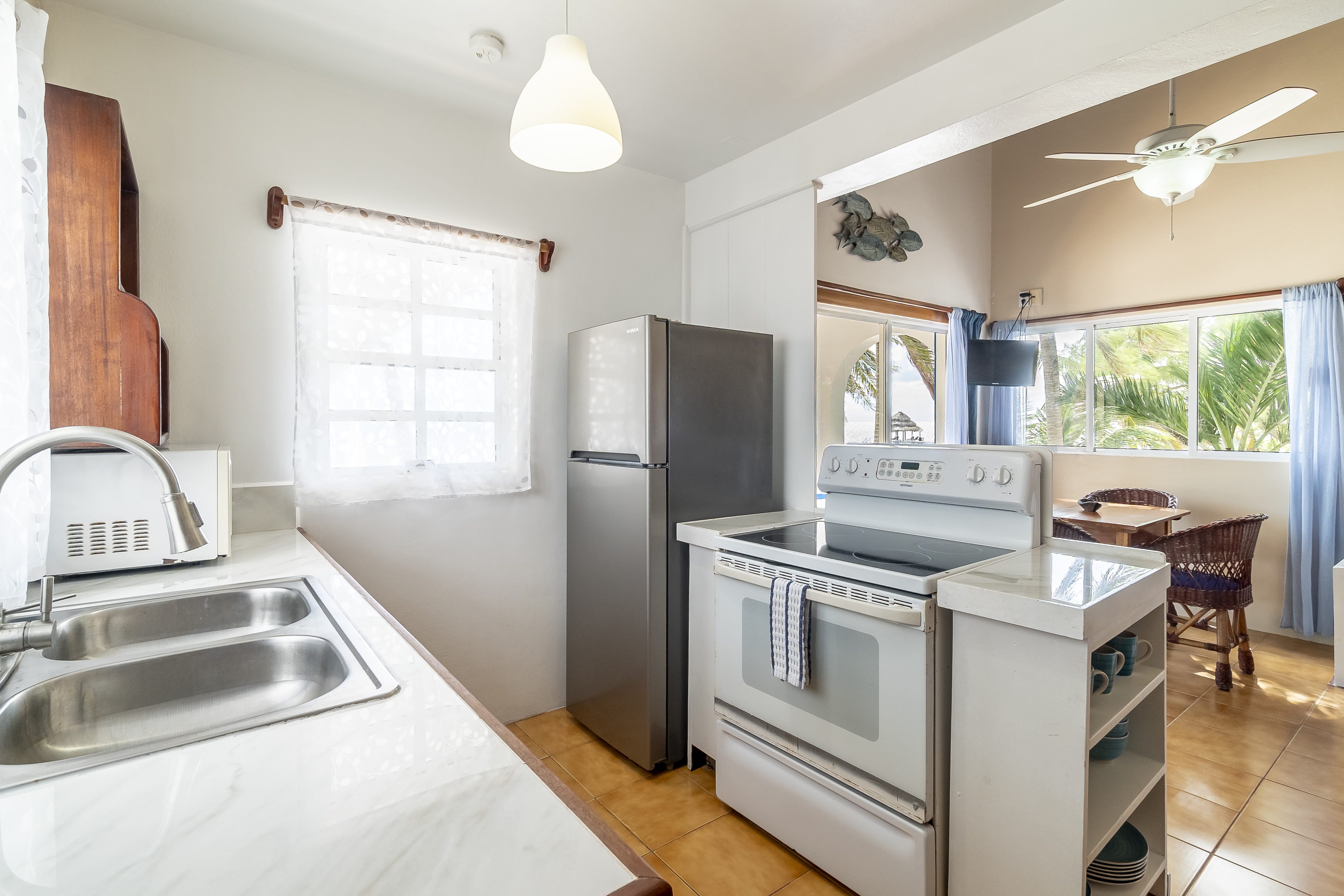 Modern, well-lit kitchen in a tropical suite featuring a stainless steel refrigerator, white electric range stove, and a white double-sink vanity. The open layout provides a clear view into the bright dining and living area.