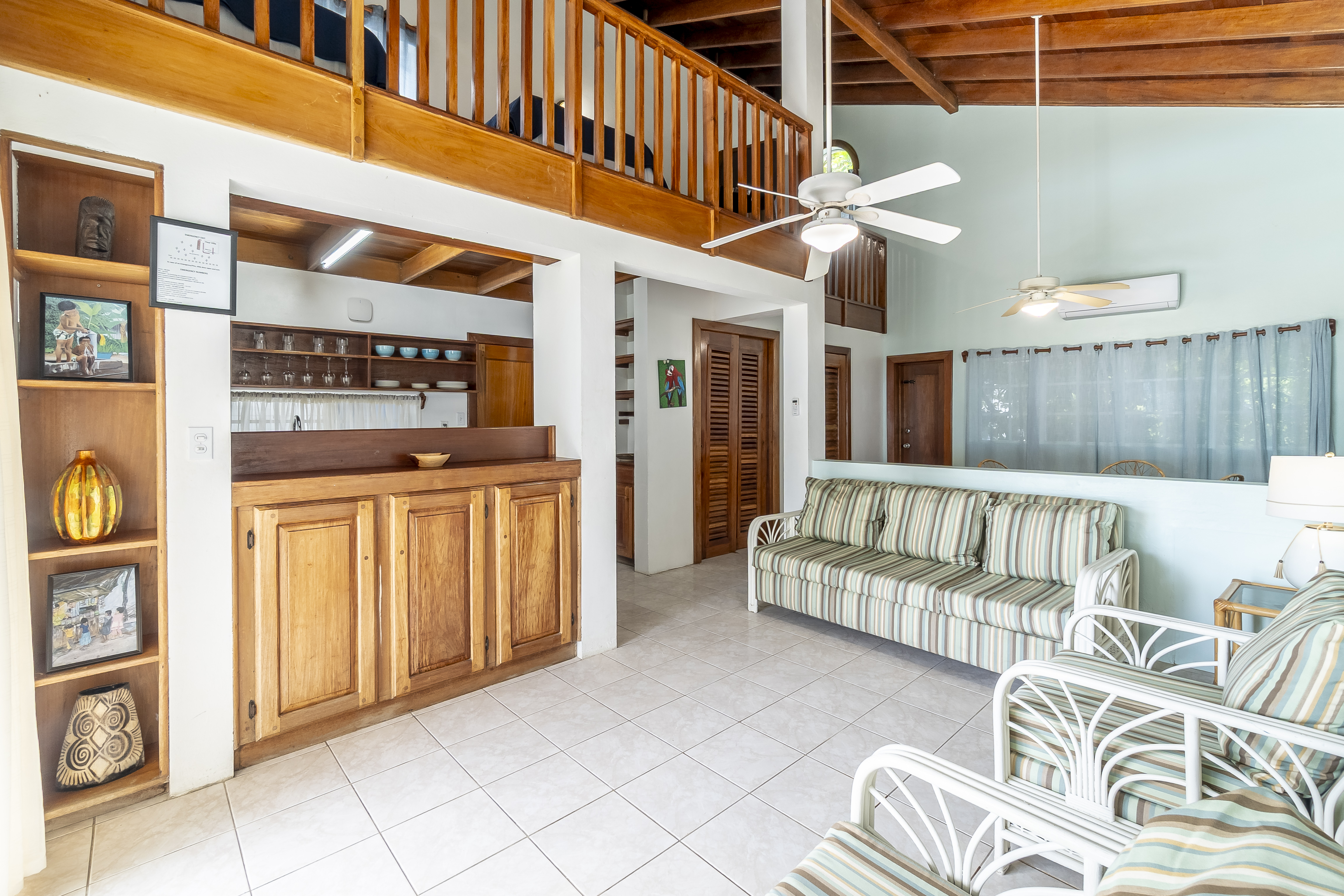 Spacious living area featuring high vaulted ceilings with wood beams, striped chairs & sofa, a breakfast bar, and a wooden balcony railing overlooking the room.