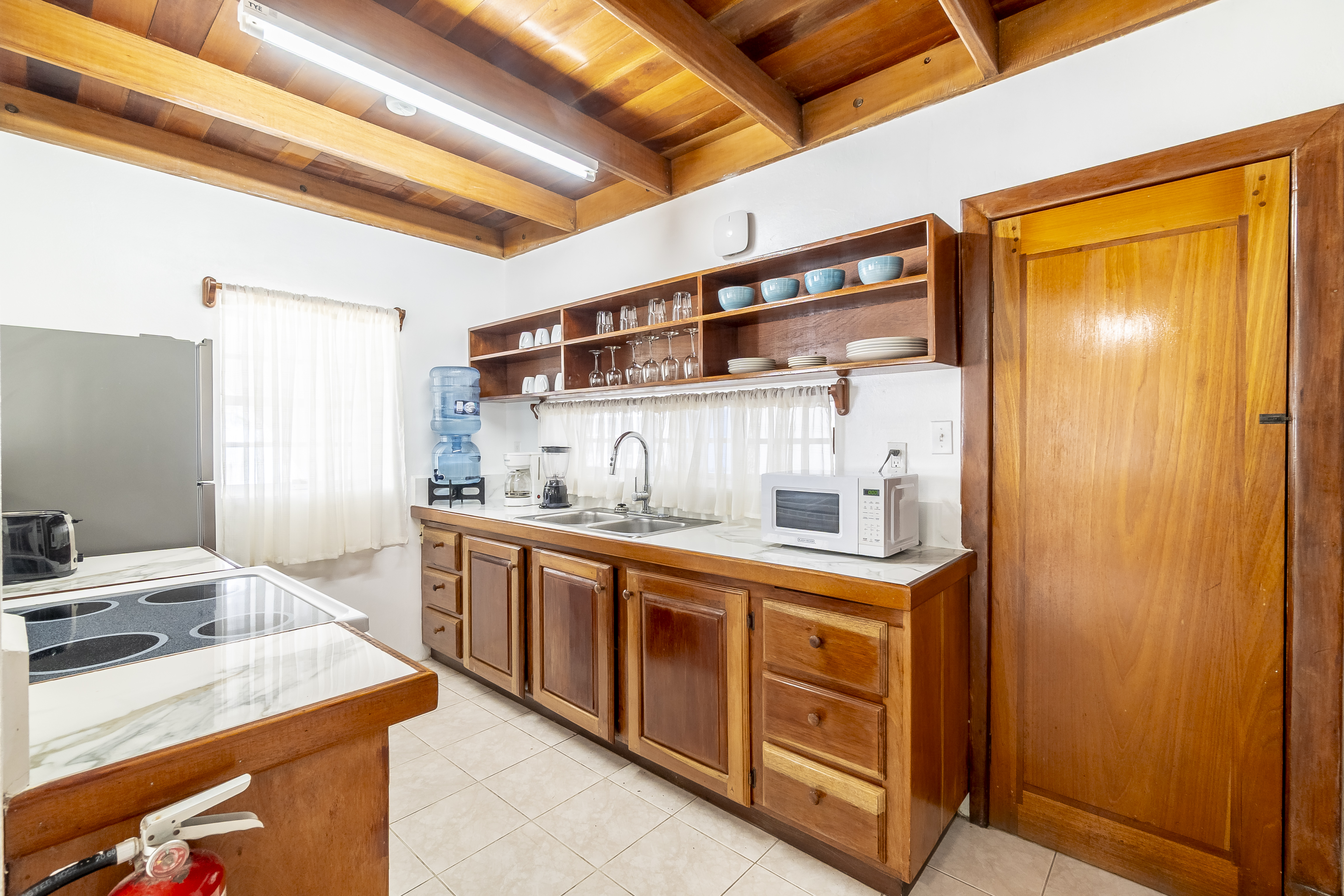 Functional kitchen in a Caribbean-style suite featuring a long countertop with a double sink, built-in wooden shelving stocked with glassware and blue dishes, a microwave, and a water dispenser, all set under a warm wood-plank ceiling.