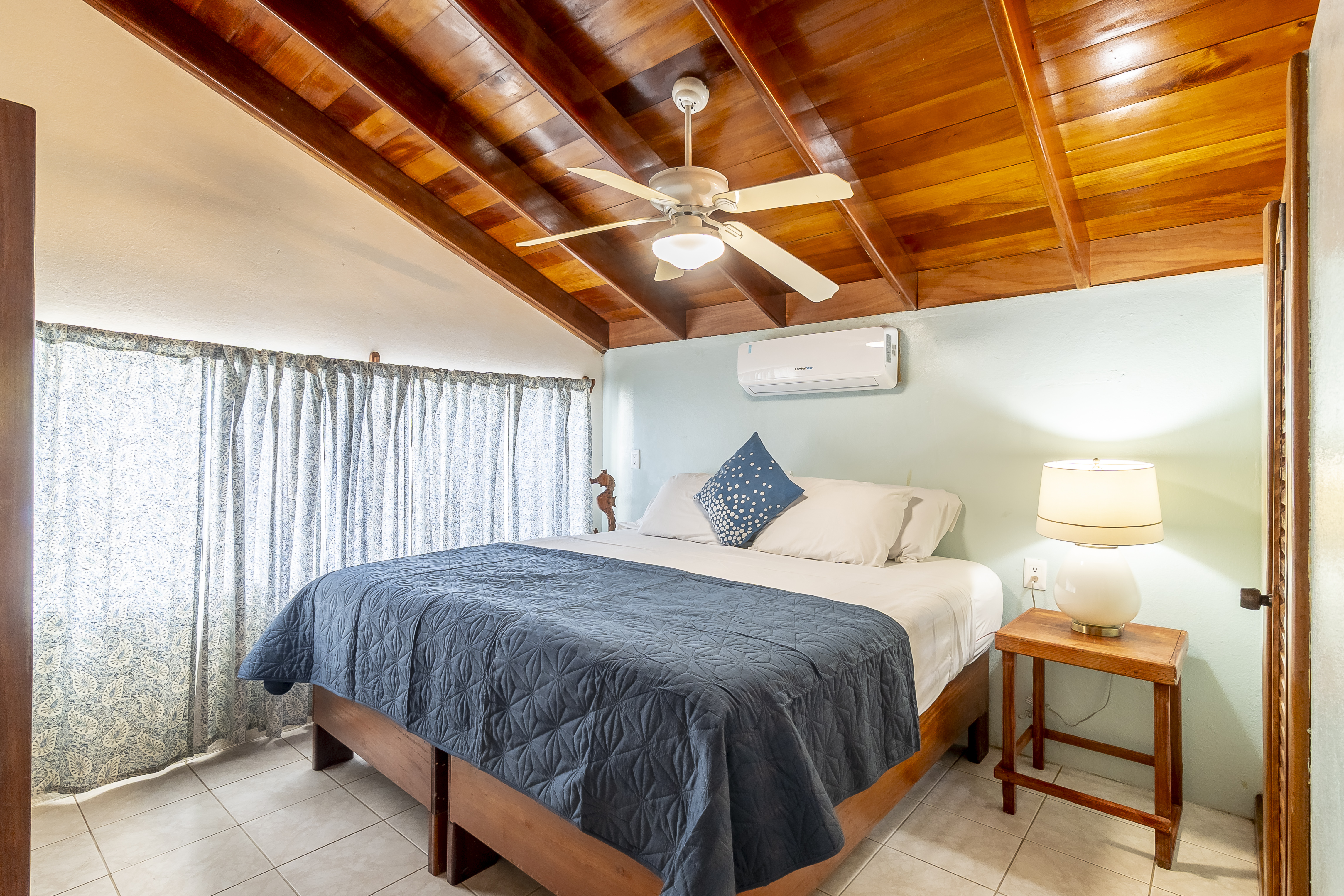 A cozy bedroom featuring a king-size bed with a dark blue coverlet, set beneath a warm, sloped wood-plank ceiling with a lit ceiling fan and an air conditioning unit.