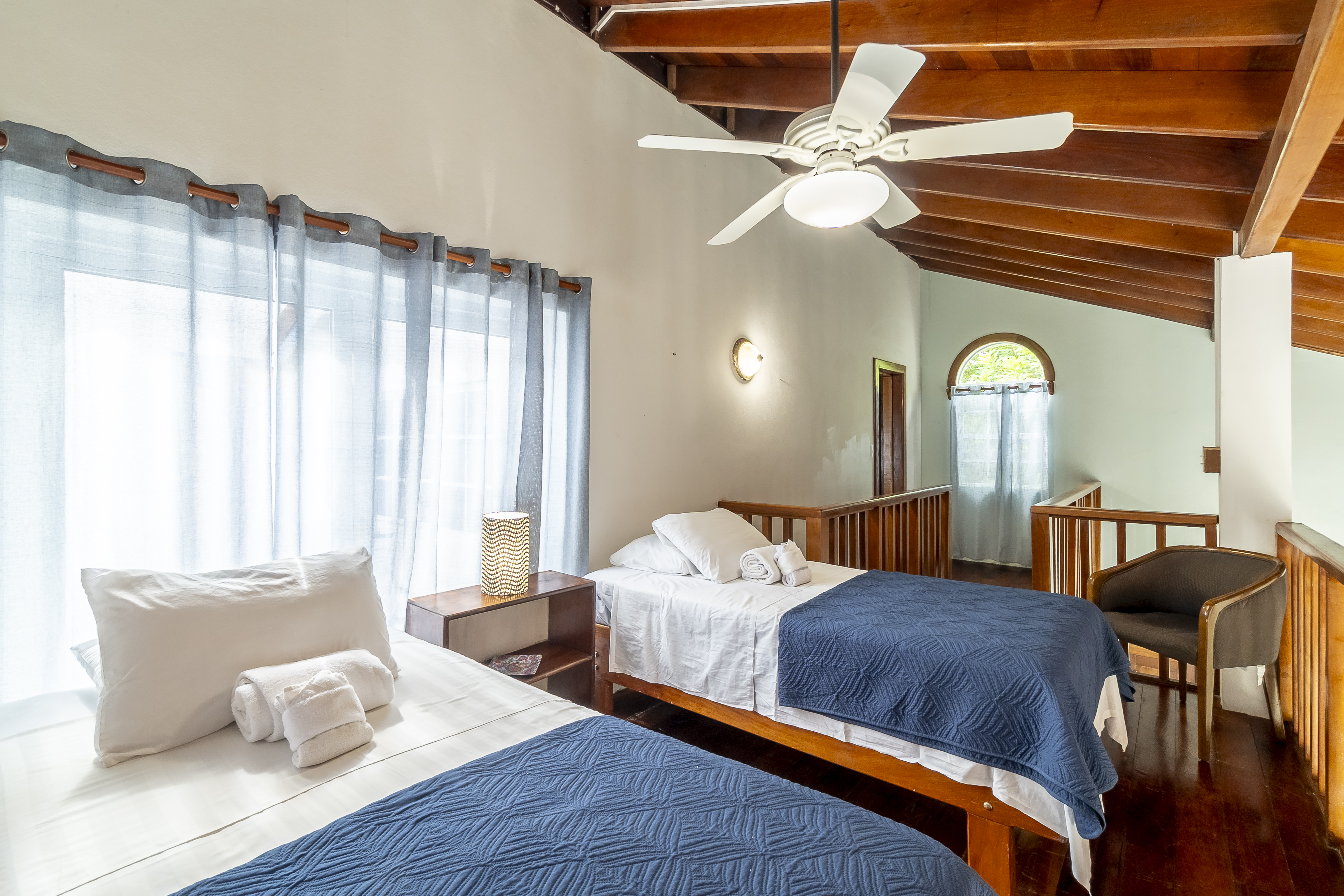 Upper-level sleeping loft featuring two twin beds with dark blue covers, polished wood flooring, a ceiling fan, and a wooden safety railing overlooking the main floor.