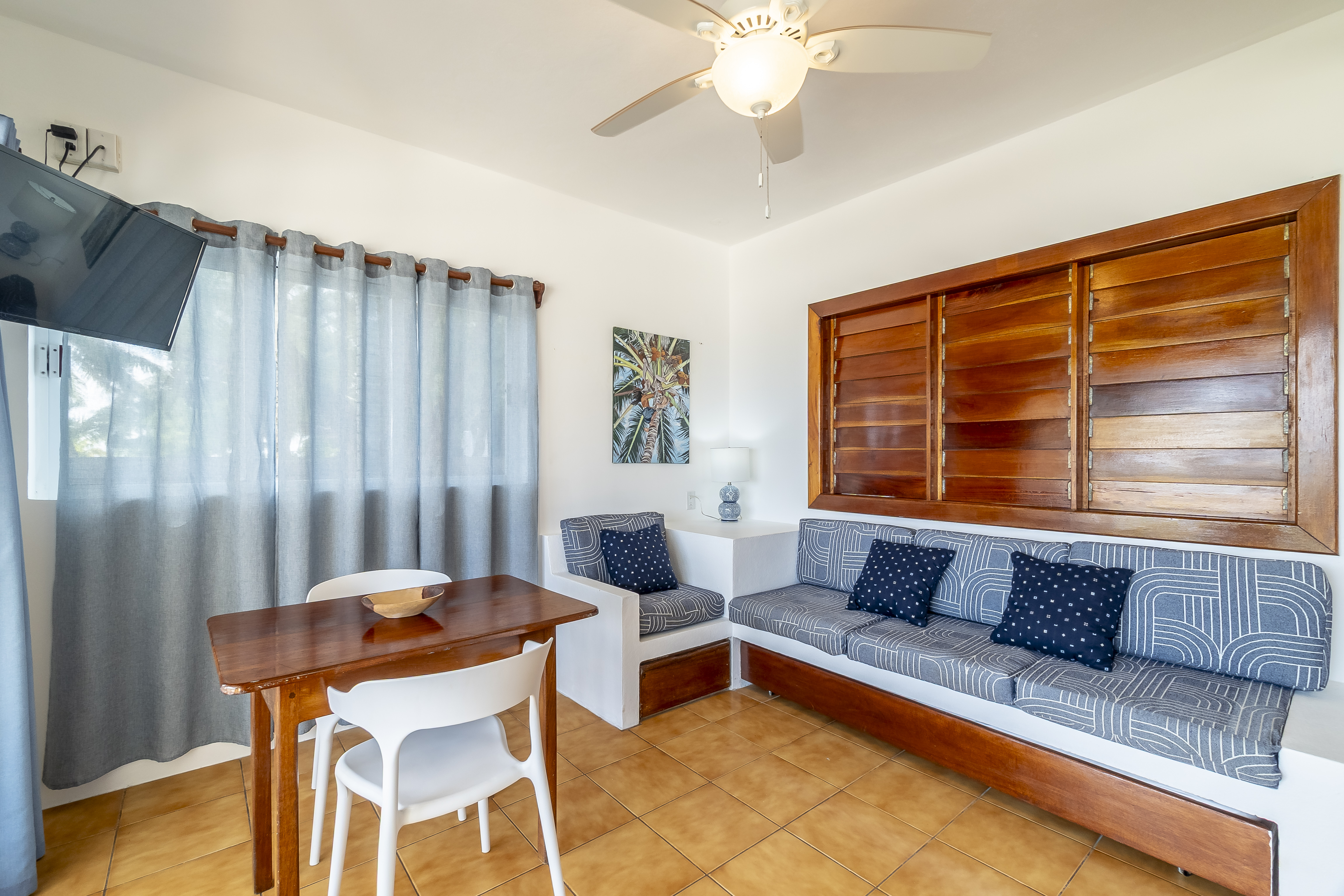 Beachfront living room in a tropical suite featuring tan floor tiles, blue and white seating, a ceiling fan, and draped large sliding glass doors.
