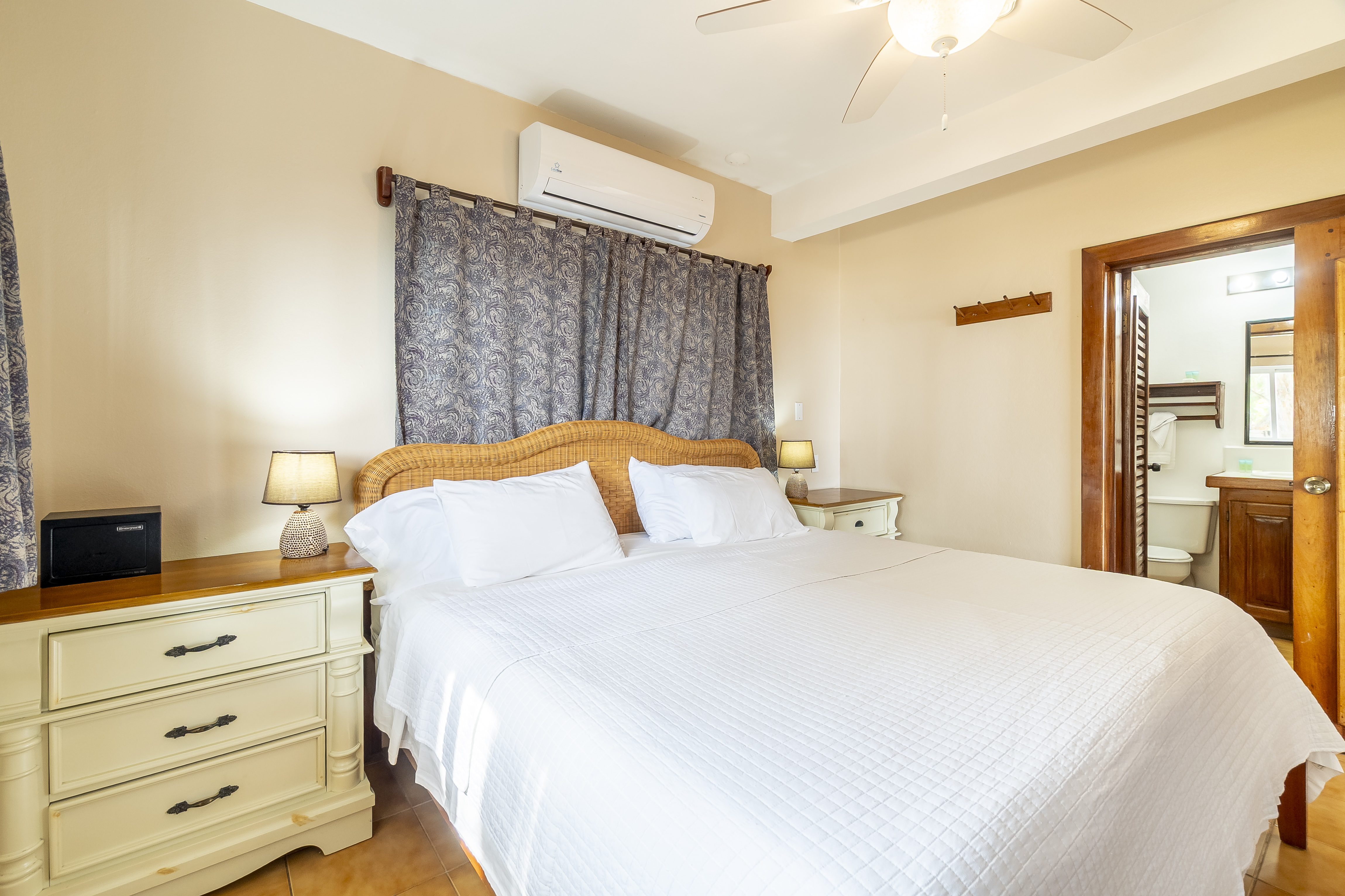 Modern bedroom in a tropical suite featuring a king-size bed with crisp white linens, a decorative wood headboard, and cream-colored bedside dressers. The room is equipped with a ceiling fan, air conditioning, and a view into an adjacent modern bathroom.
