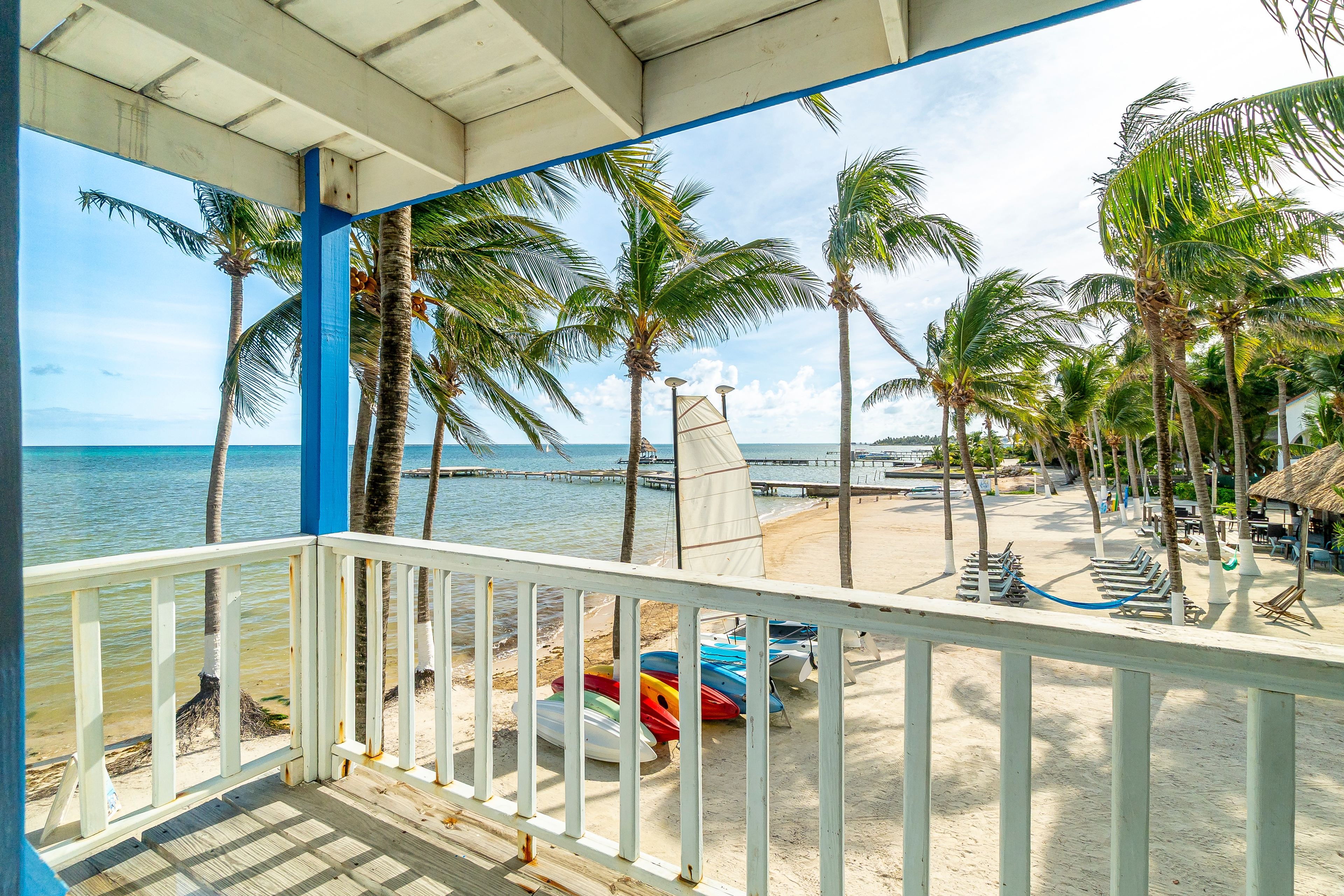 Beachfront balcony view overlooking a white sand beach with palm trees, colorful kayaks, and a sailboat, with the calm blue Caribbean Sea in the background.