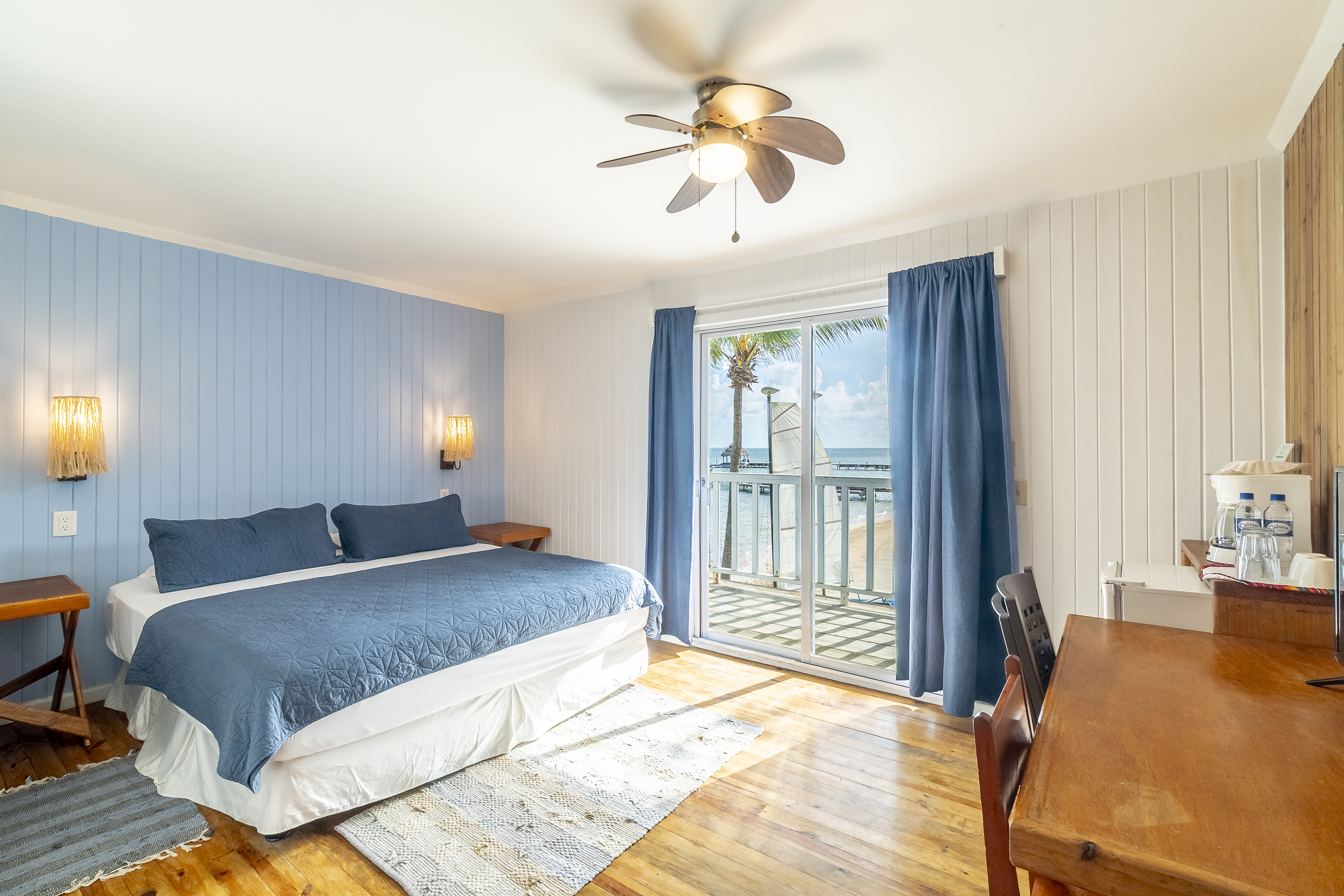 Modern beachfront bedroom featuring a king-size bed with a navy blue quilt, light blue wood-paneled walls, and large glass sliding doors that open to a balcony with views of the Caribbean Sea and palm trees.