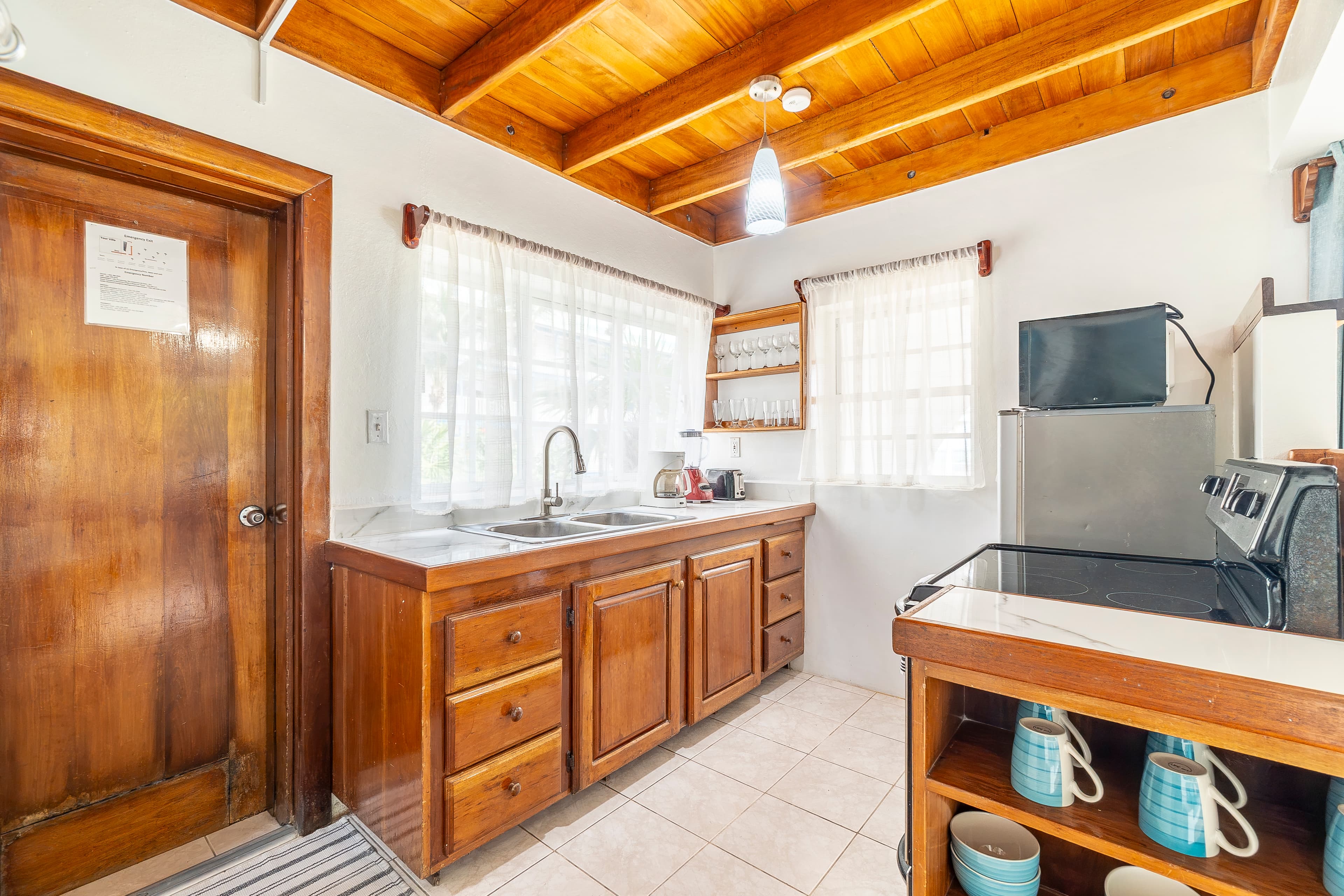 Brightly lit kitchen area featuring warm wood cabinetry, a white countertop with a double sink, and a high wood-plank ceiling. The space includes a stove, microwave, and refrigerator.