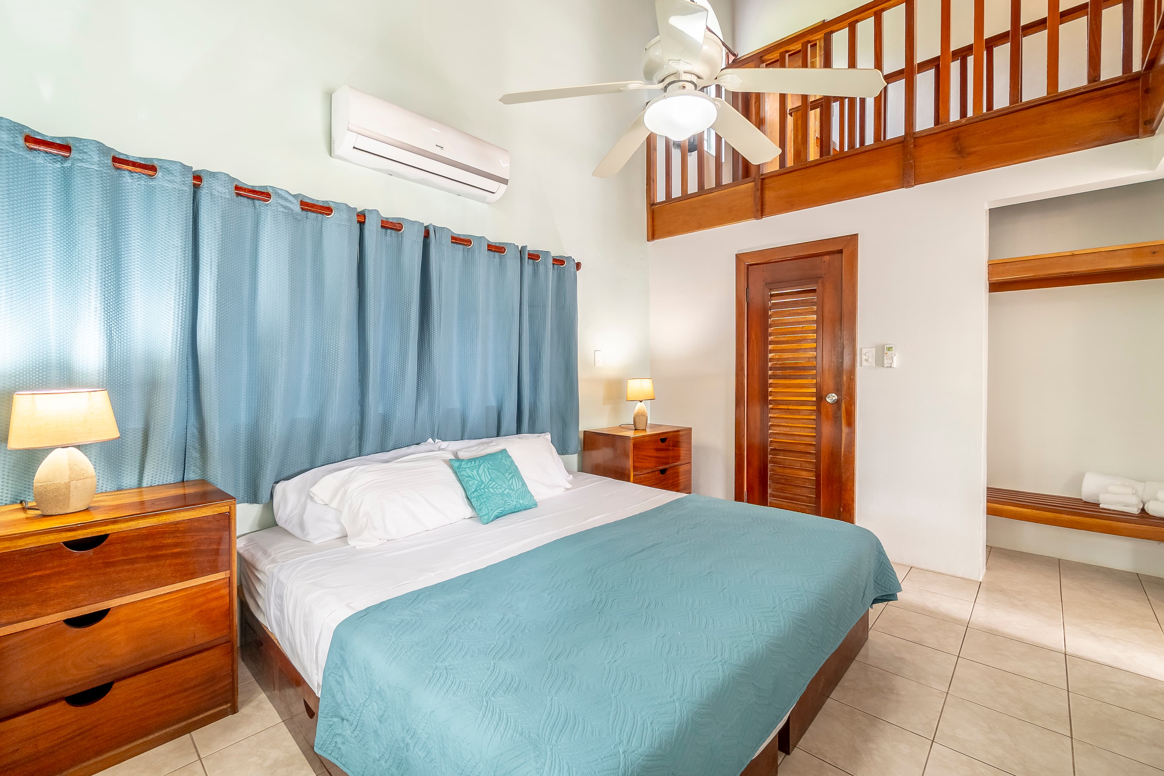A brightly lit master bedroom featuring a king-size bed with light blue bedding, warm wood furniture, air conditioning, and a wooden loft railing overlooking the space from above.