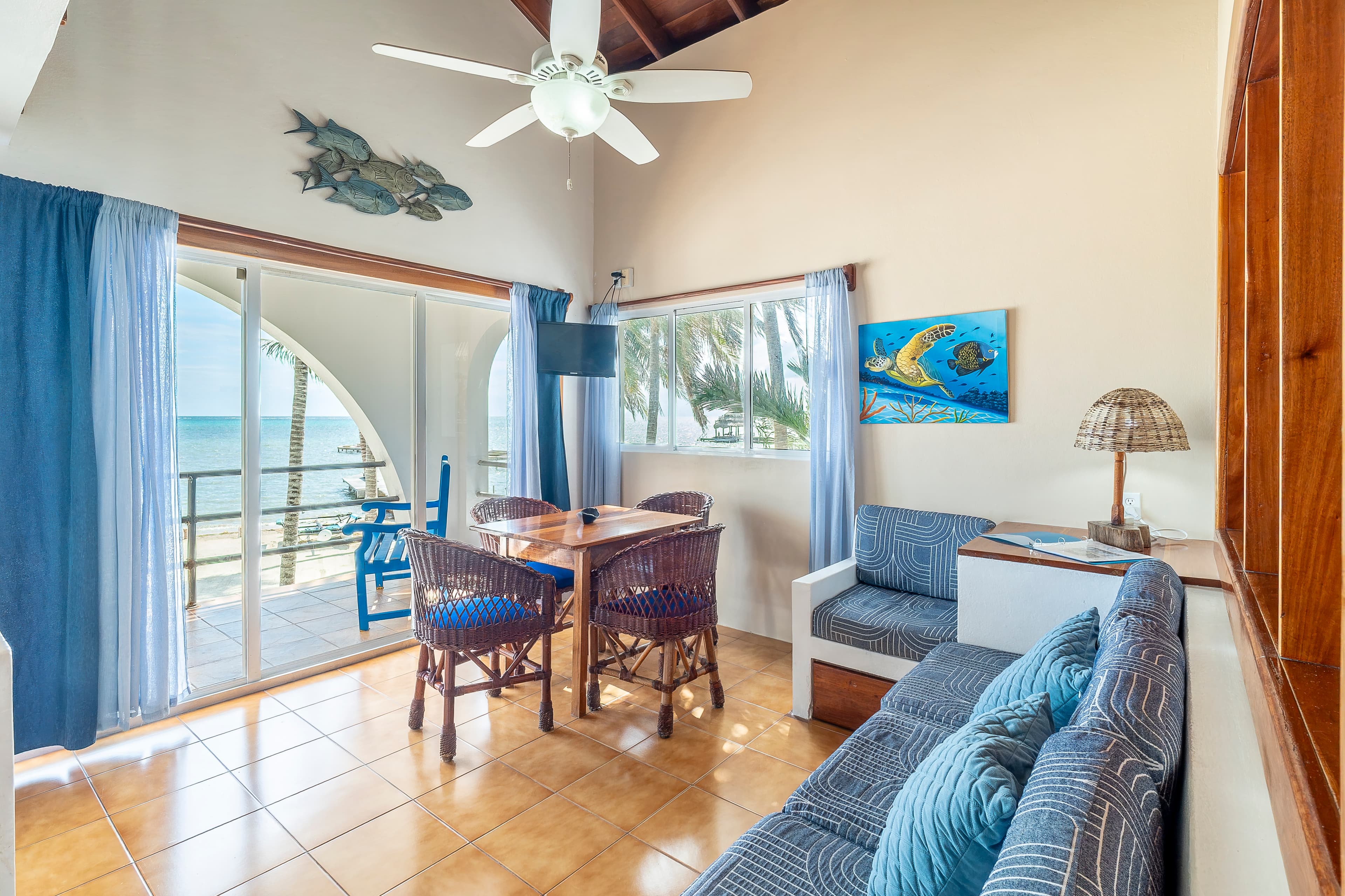 Brightly lit living room featuring light blue sofas, a wooden dining set, and sliding glass doors that open to a balcony with a stunning view of the turquoise Caribbean Sea.