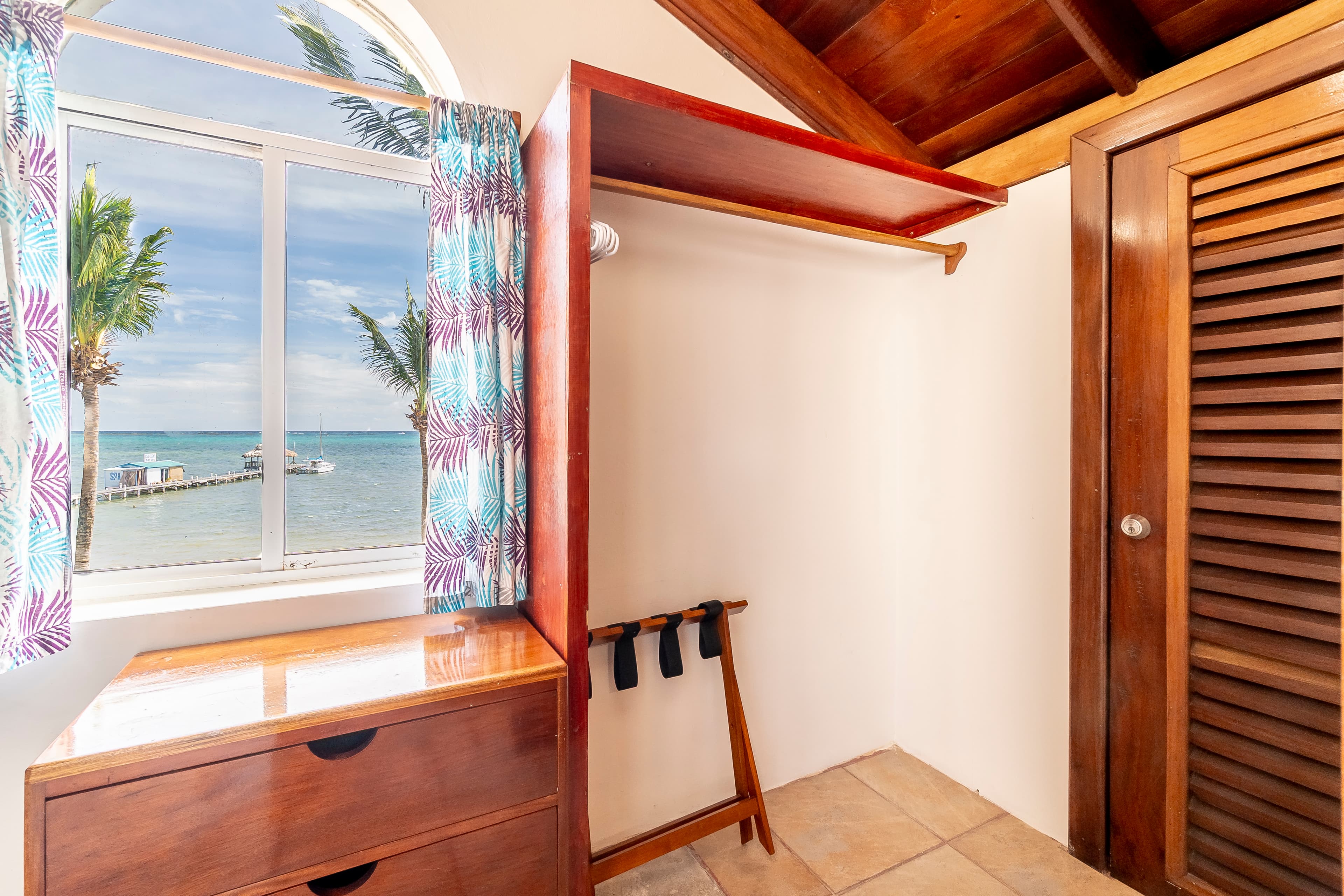 Clean, white-tiled bathroom in a tropical suite featuring a wooden vanity with a white countertop and sink, a large mirror with a warm wooden frame, and a white toilet.