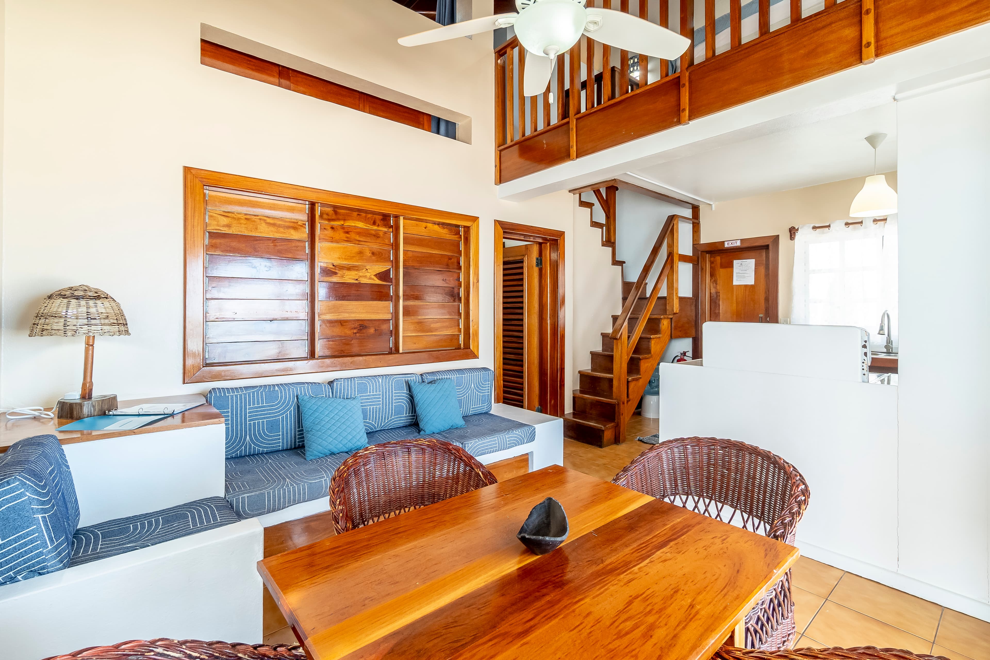 Open-concept living space in a tropical suite featuring a wooden dining table with wicker chairs, a built-in blue sofa, a ceiling fan, and a wooden staircase leading to an upper-level loft.