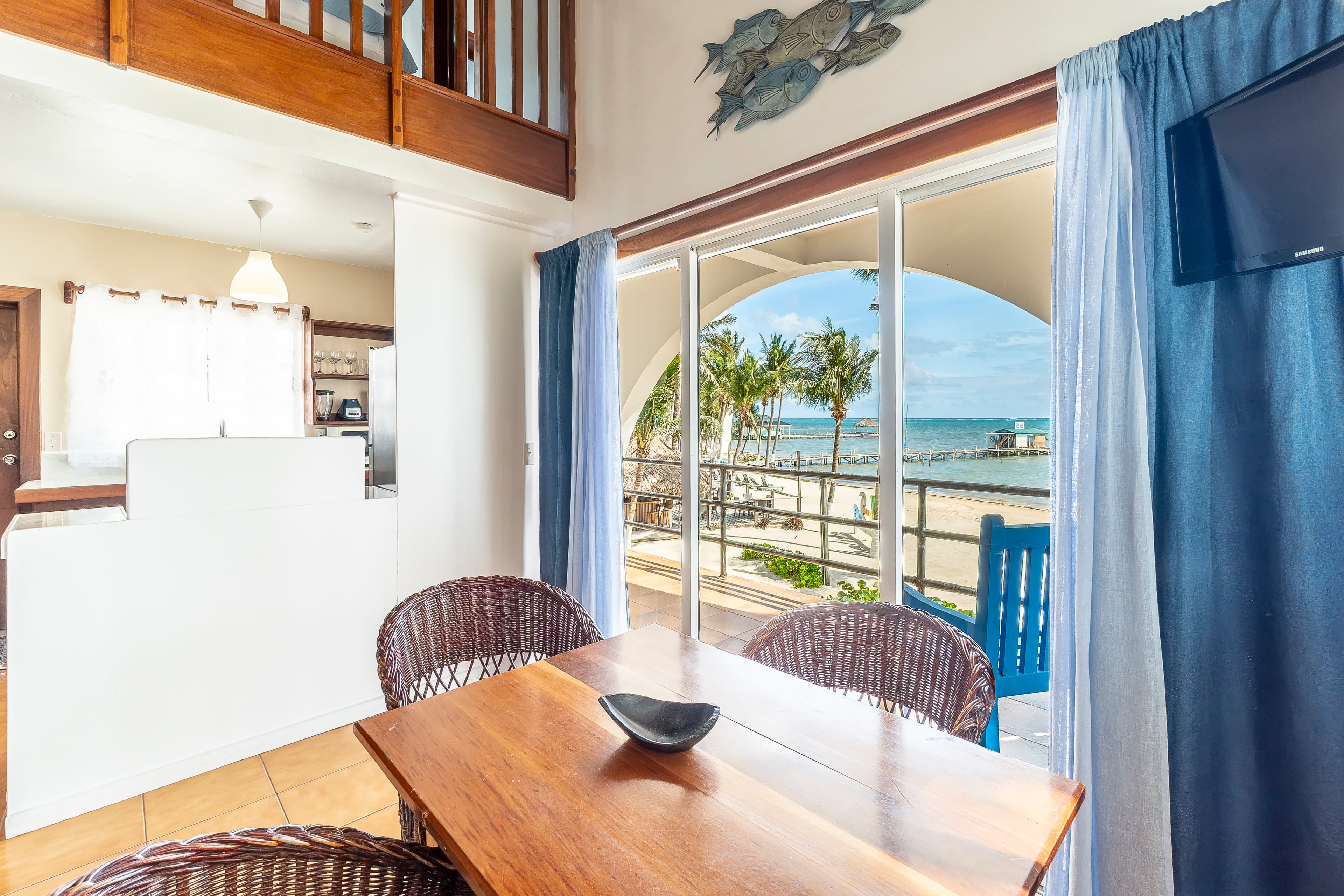 Beachfront dining area inside a tropical suite featuring a wooden table with wicker chairs, situated next to large sliding glass doors that offer a clear view of the sandy beach and blue Caribbean Sea.
