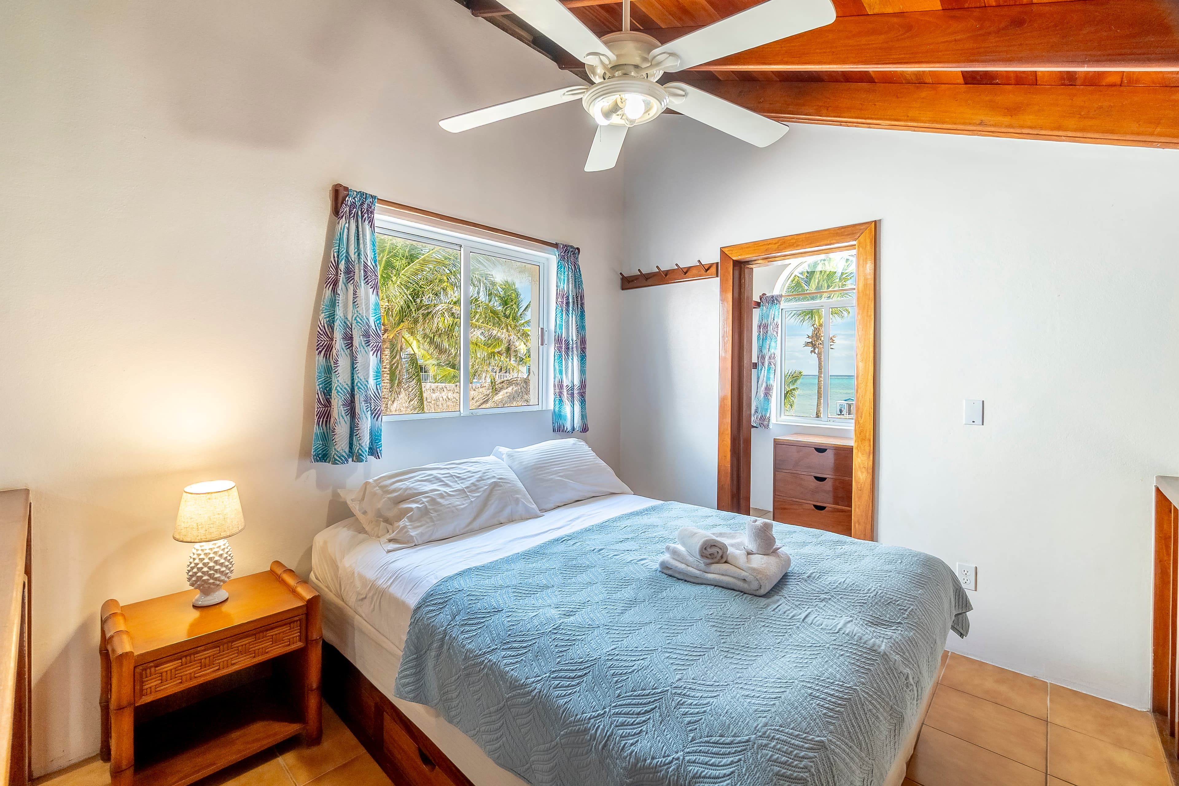A cozy bedroom in a tropical suite featuring a queen-size bed with light blue bedding, set beneath a vaulted wood-plank ceiling with a lit ceiling fan and an air conditioning unit.