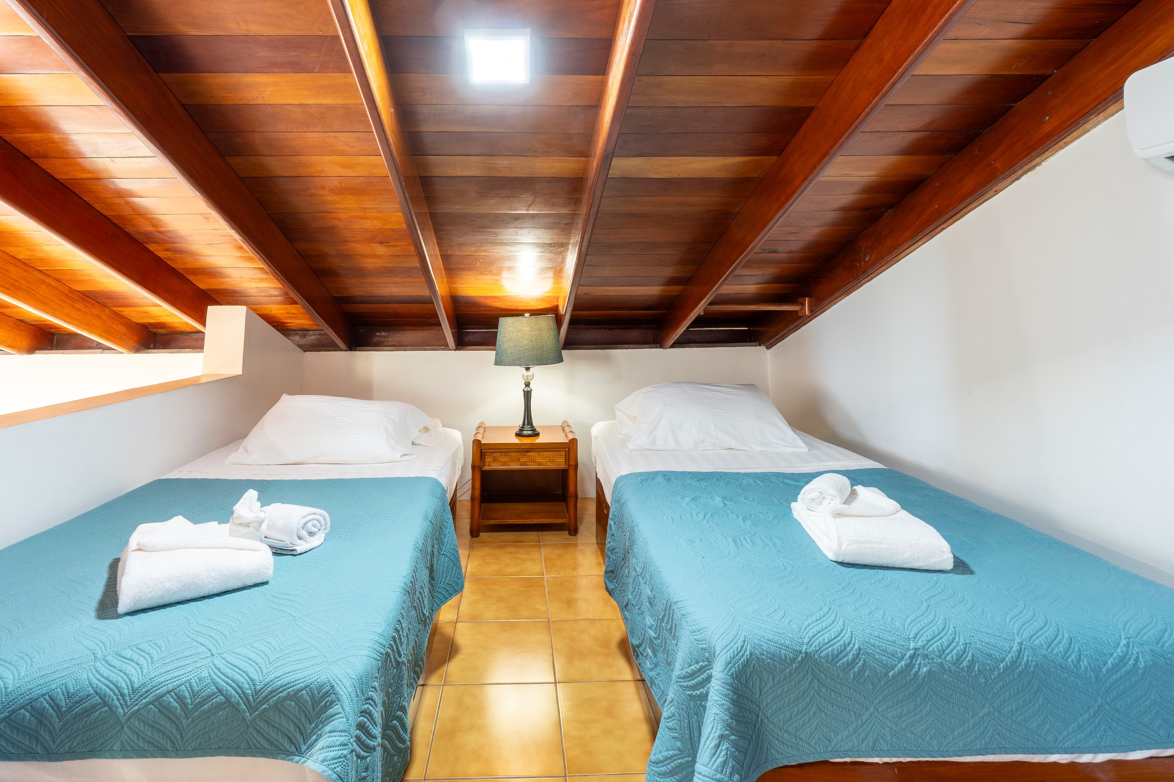 Upper-level sleeping loft with two twin beds featuring teal quilted bedding, located under a sloped wooden ceiling with a central nightstand and lamp.