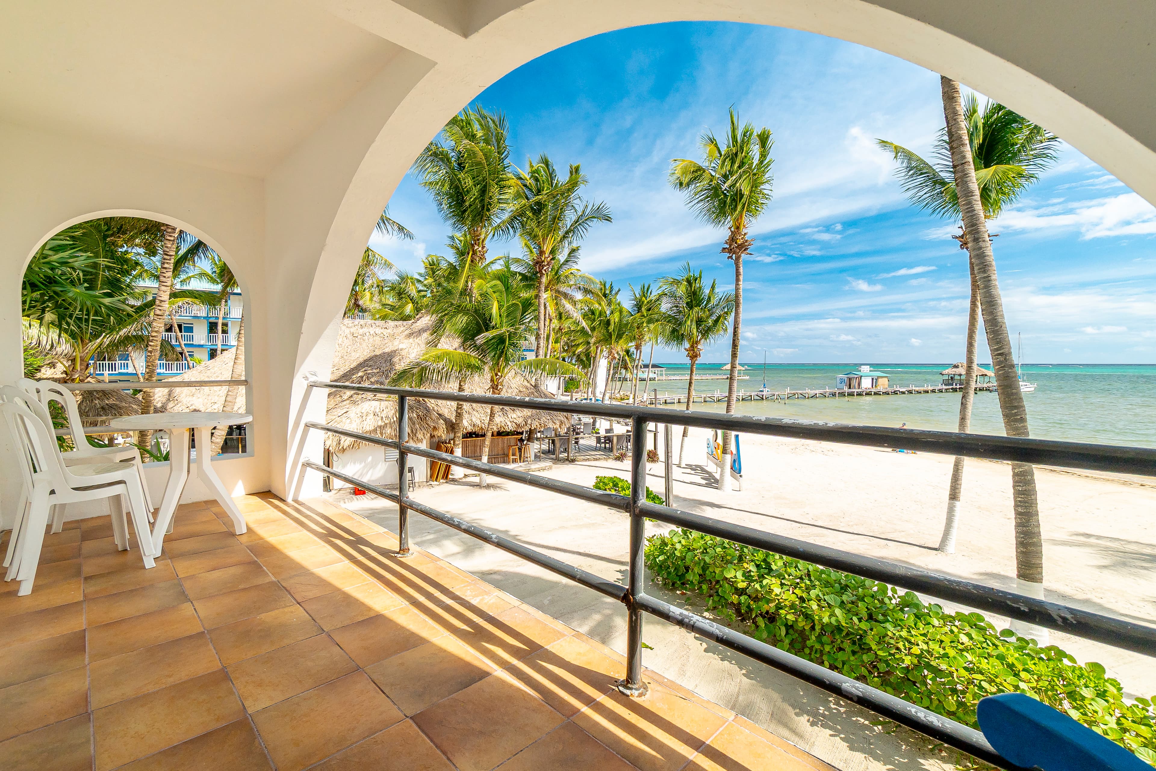 View from a spacious private balcony through a white arched opening, featuring a small patio table and chairs, overlooking a palm-lined white sand beach and the turquoise Caribbean Sea.