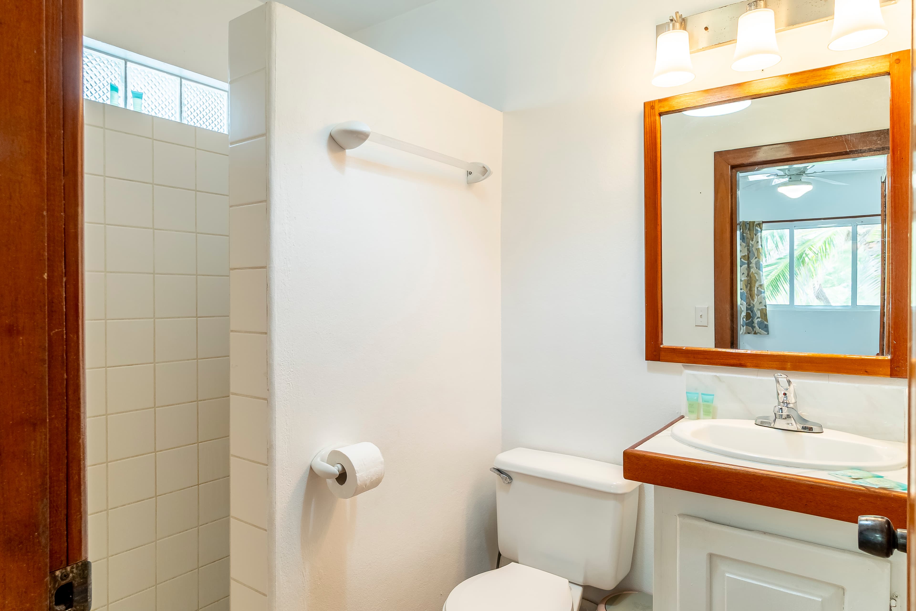 Modern bathroom in a tropical suite featuring a wooden vanity with a white countertop and sink, a large mirror with warm vanity lighting, a toilet, and a white-tiled walk-in shower area.