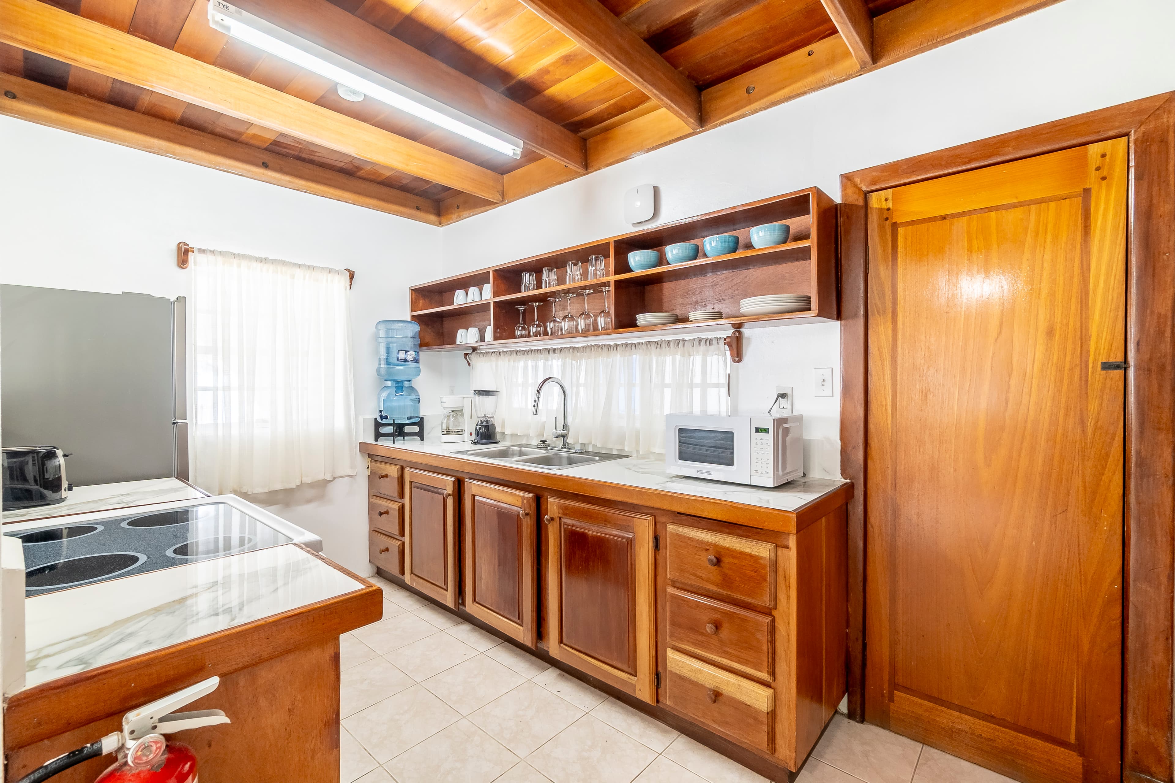 Functional kitchen in a Caribbean-style suite featuring a long countertop with a double sink, built-in wooden shelving stocked with glassware and blue dishes, a microwave, and a water dispenser, all set under a warm wood-plank ceiling.