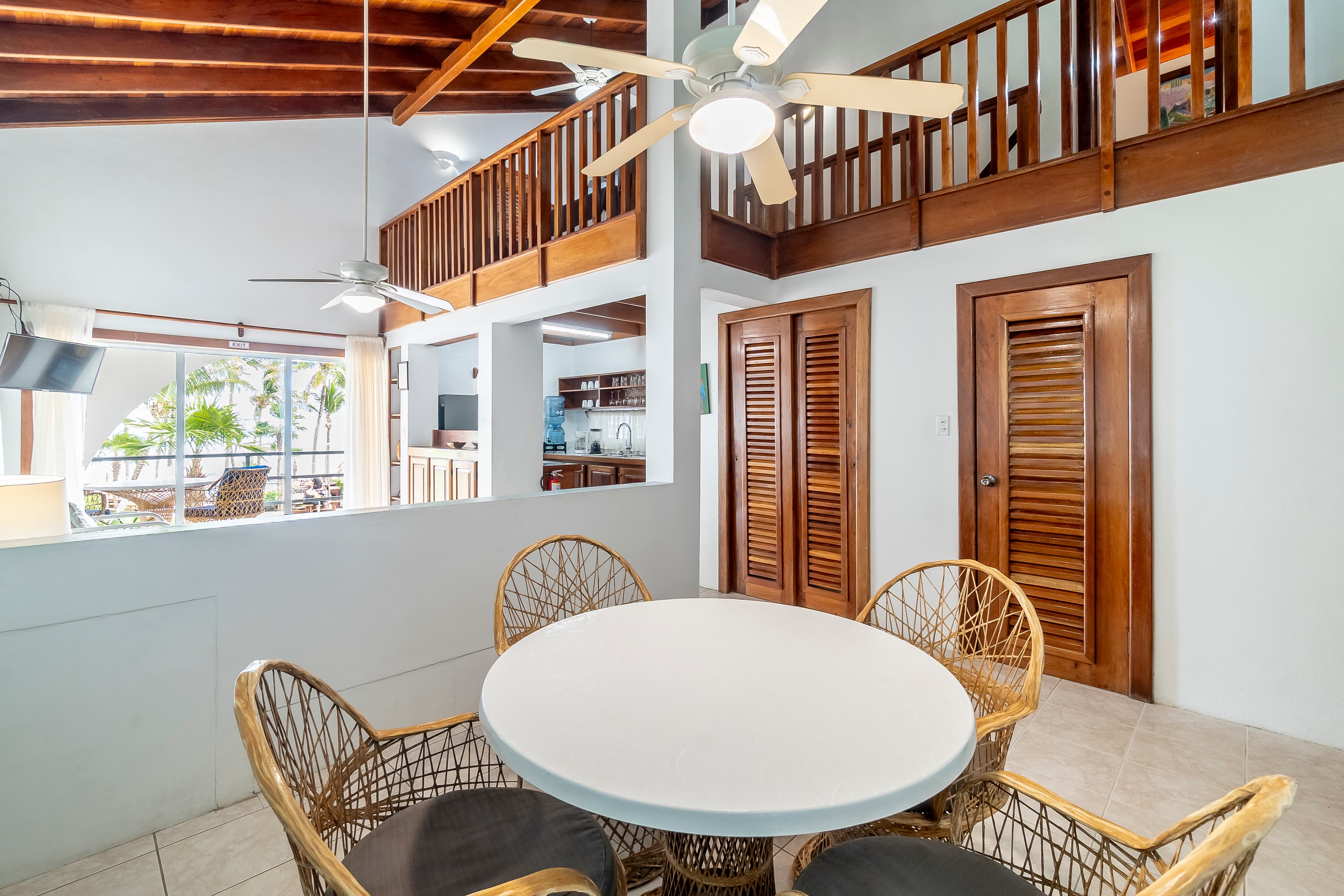 A bright dining area in a tropical suite featuring a round white table with four wicker chairs, set against a background of high vaulted wood-beam ceilings and a second-level wooden loft railing.