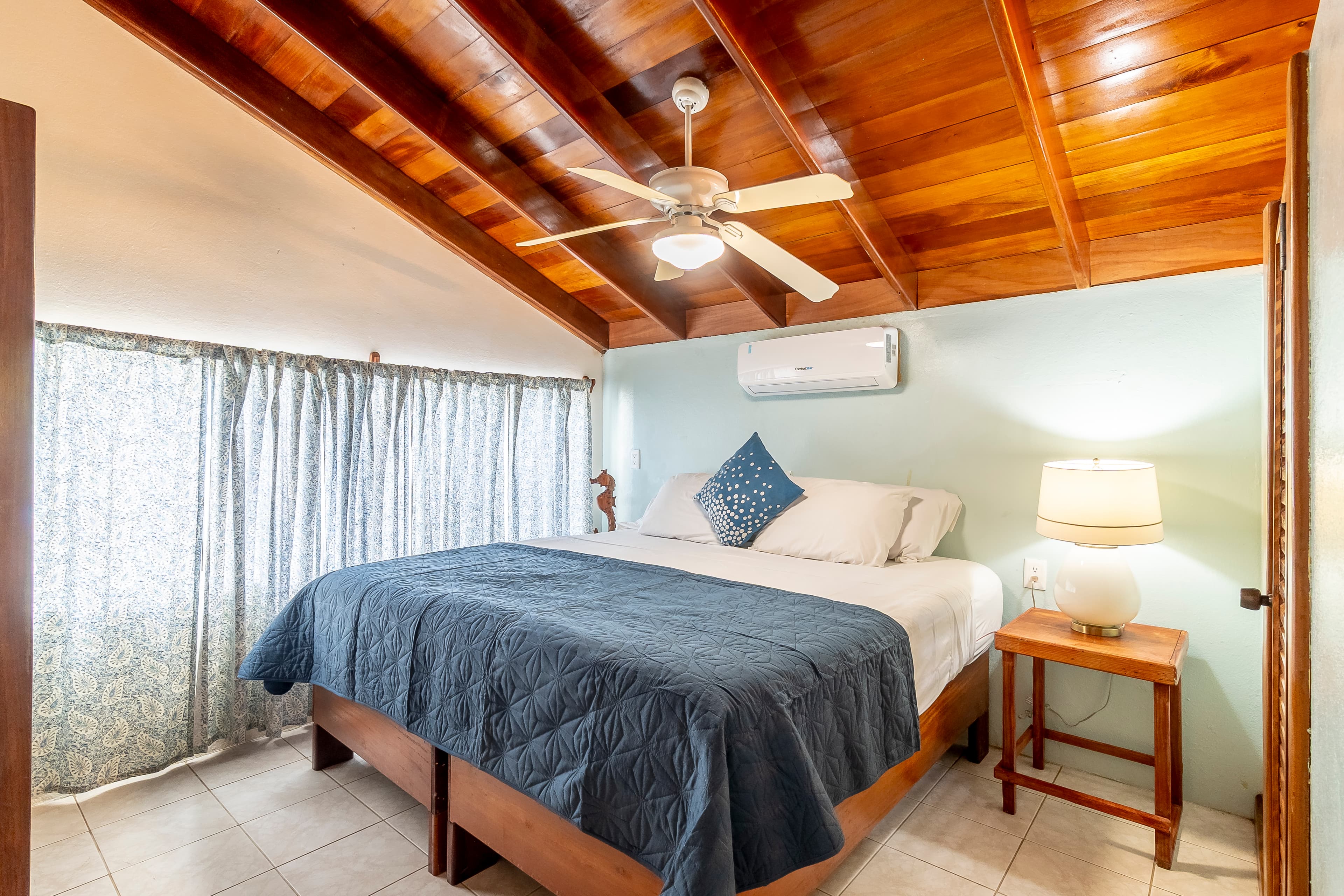 A cozy bedroom featuring a king-size bed with a dark blue coverlet, set beneath a warm, sloped wood-plank ceiling with a lit ceiling fan and an air conditioning unit.