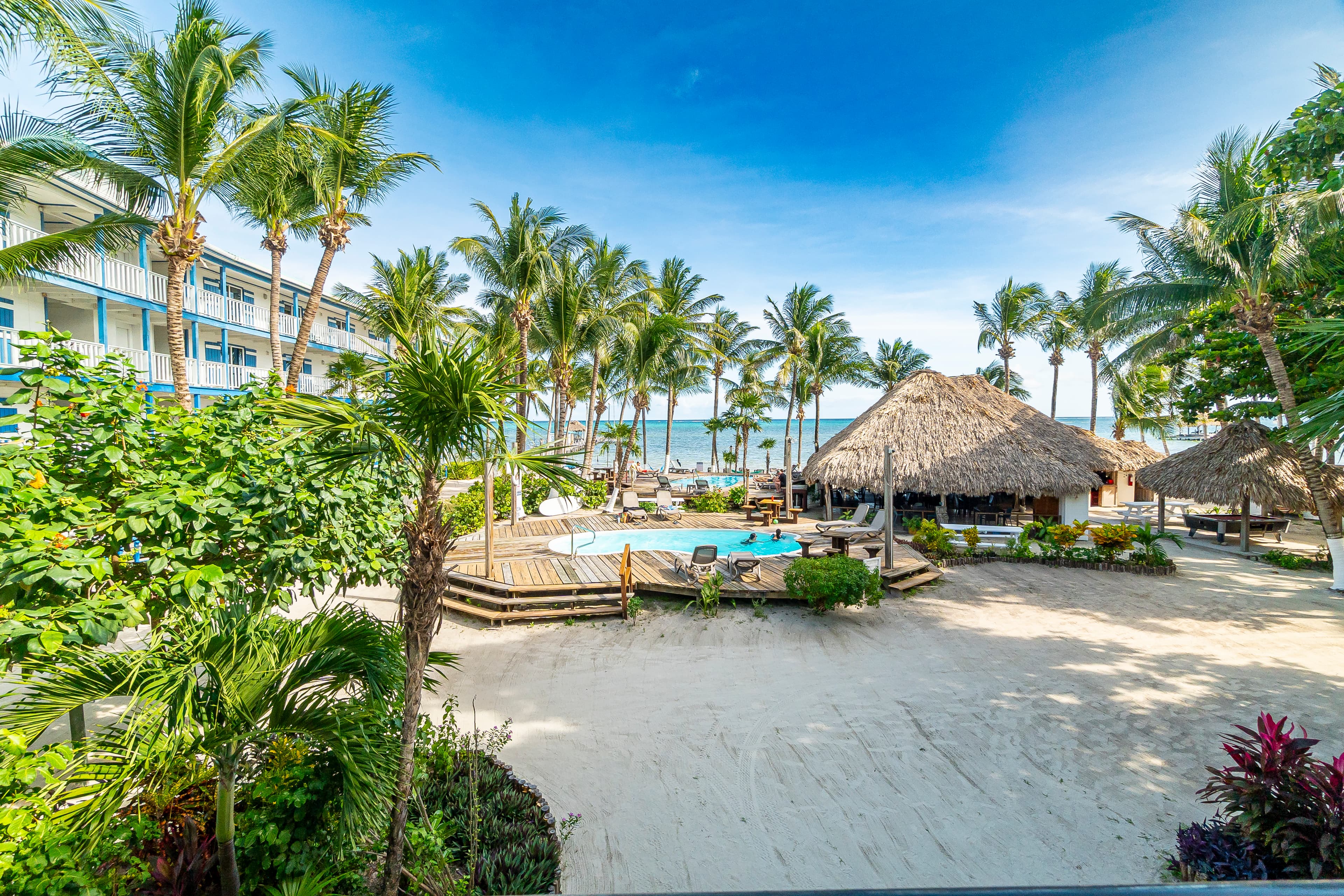 View from veranda of a tropical resort featuring a white sand courtyard, a swimming pool surrounded by palm trees, and a thatched-roof palapa bar with a glimpse of the blue Caribbean Sea in the background.