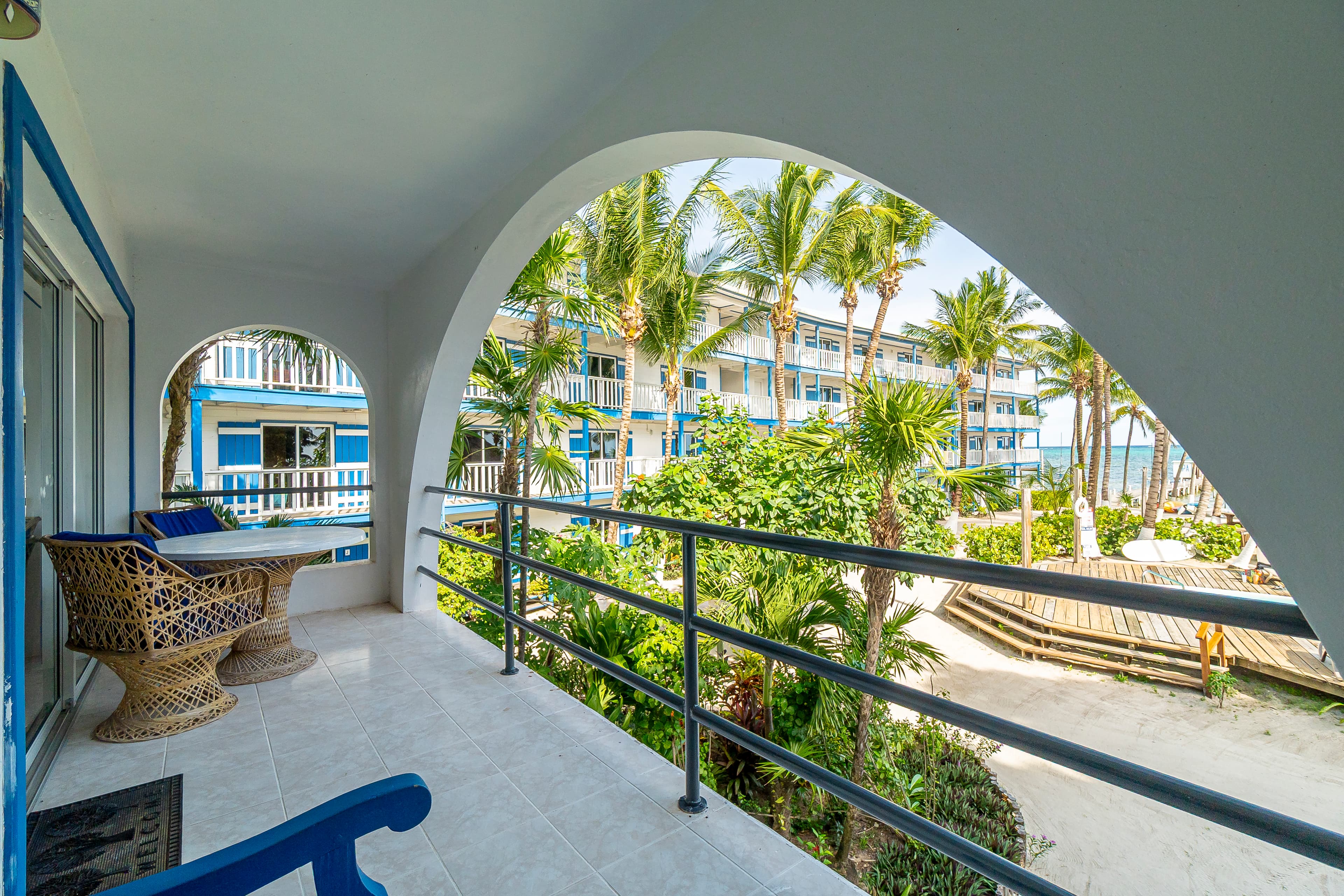 View from a private white-tiled veranda with an arched opening and metal railing, overlooking a tropical resort courtyard filled with palm trees, white sand, and beachfront buildings.
