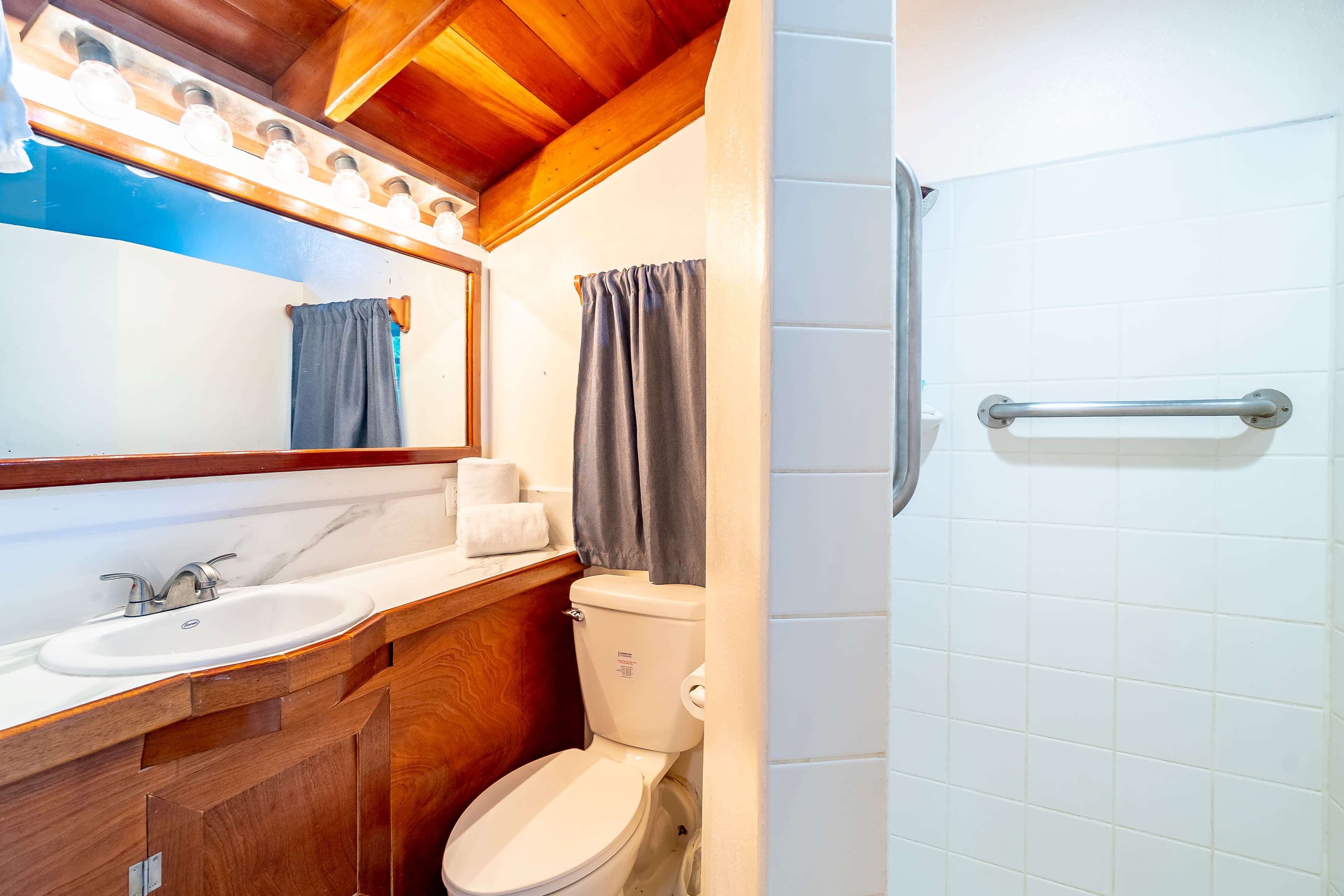 Modern bathroom in a tropical suite featuring a wooden vanity with a white countertop, a large mirror with theater-style lighting, a toilet, and a white-tiled walk-in shower with a safety grab bar.