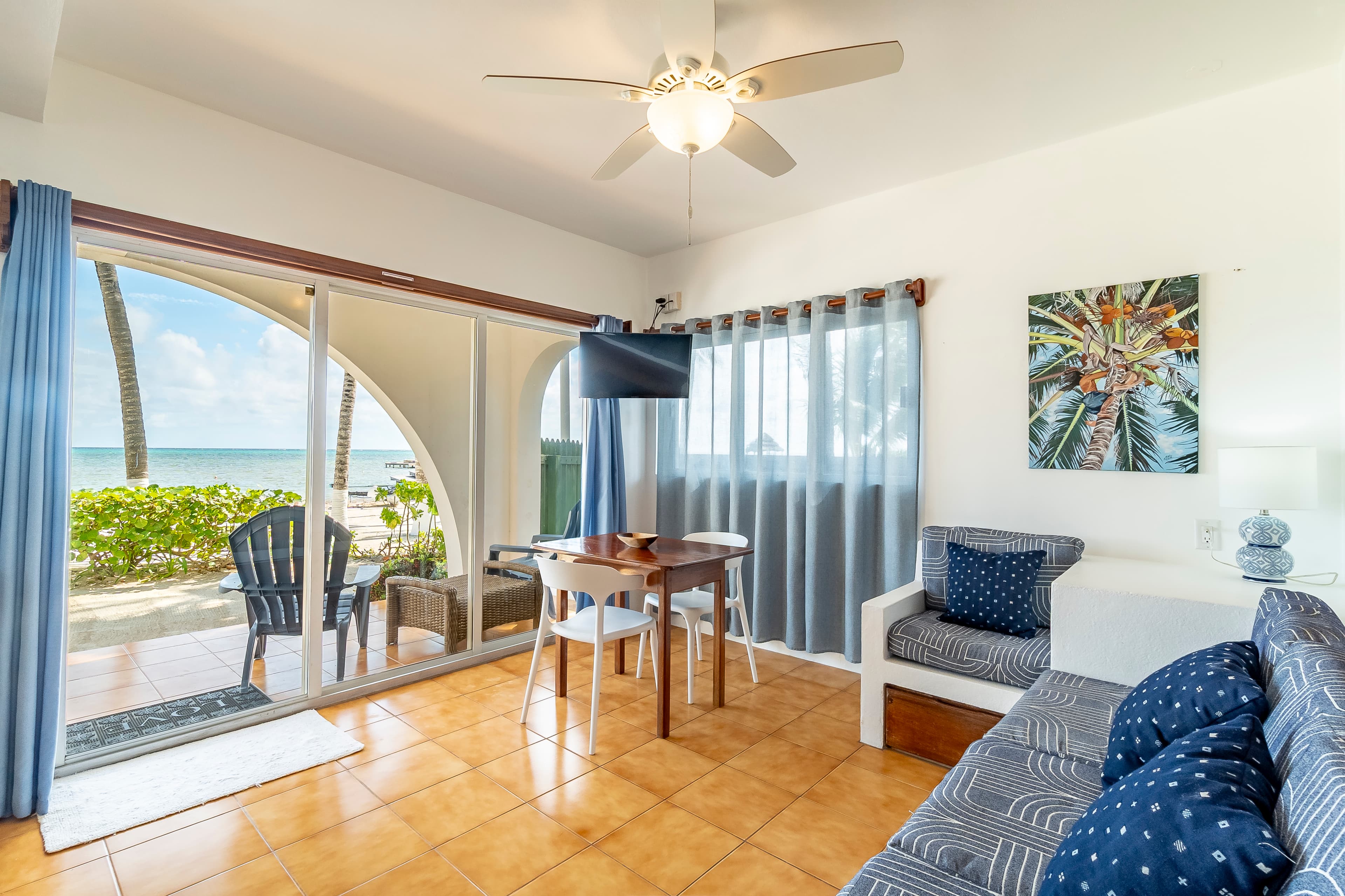 Beachfront living room in a tropical suite featuring tan floor tiles, blue and white seating, a ceiling fan, and large sliding glass doors that open to a private balcony with a view of the Caribbean Sea and lush palm trees.