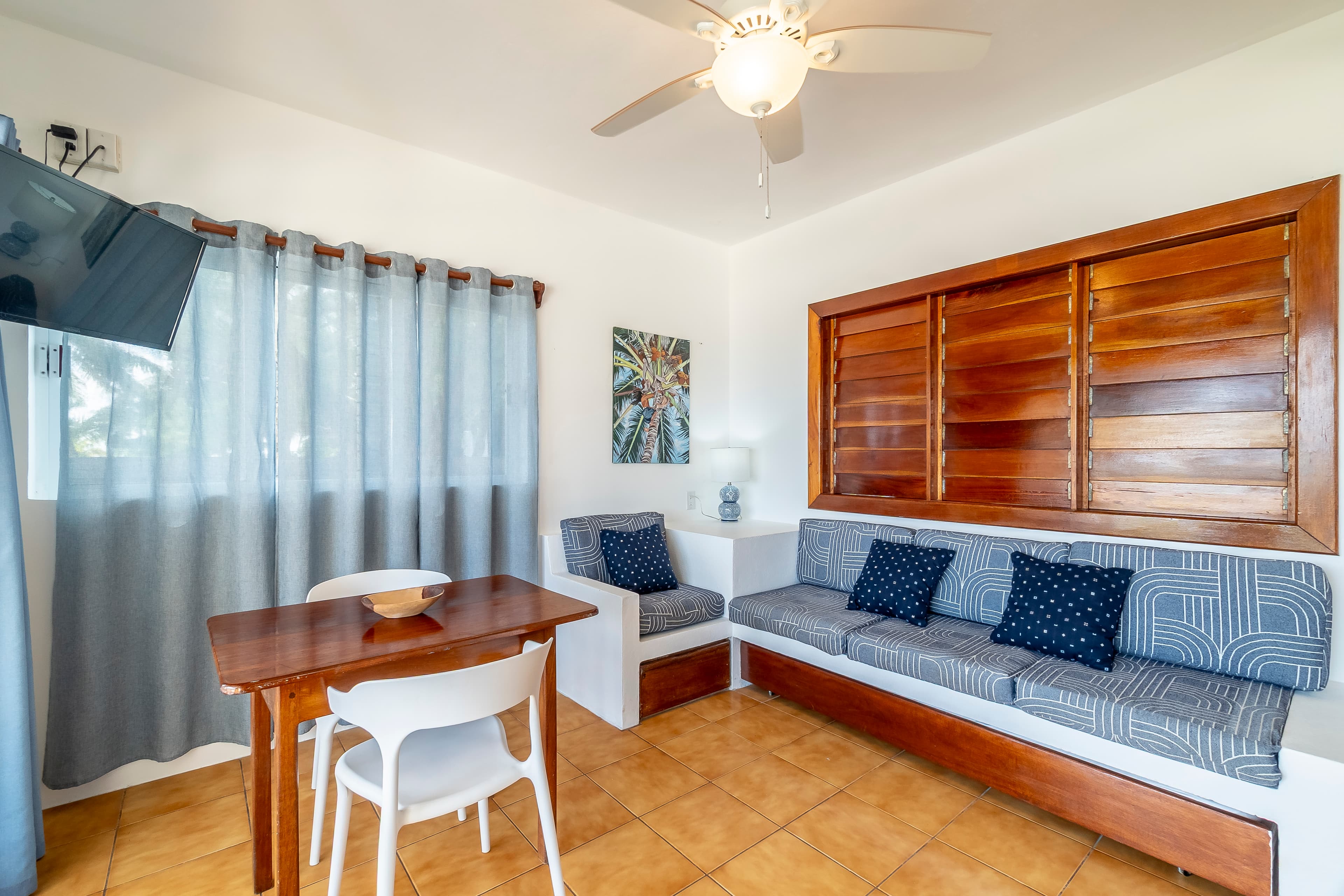 Beachfront living room in a tropical suite featuring tan floor tiles, blue and white seating, a ceiling fan, and draped large sliding glass doors.