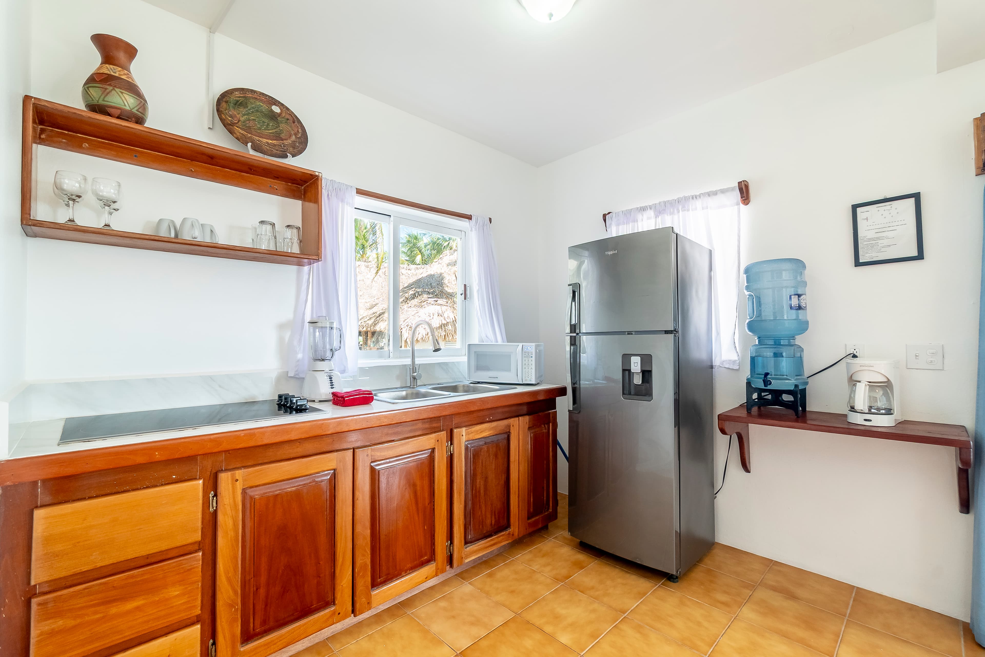 Modern kitchen area in a tropical suite featuring warm wood cabinetry, a stainless steel refrigerator, and a water dispenser. The space includes a white countertop with a double sink, built-in wooden shelving for glassware, and a window overlooking lush greenery.