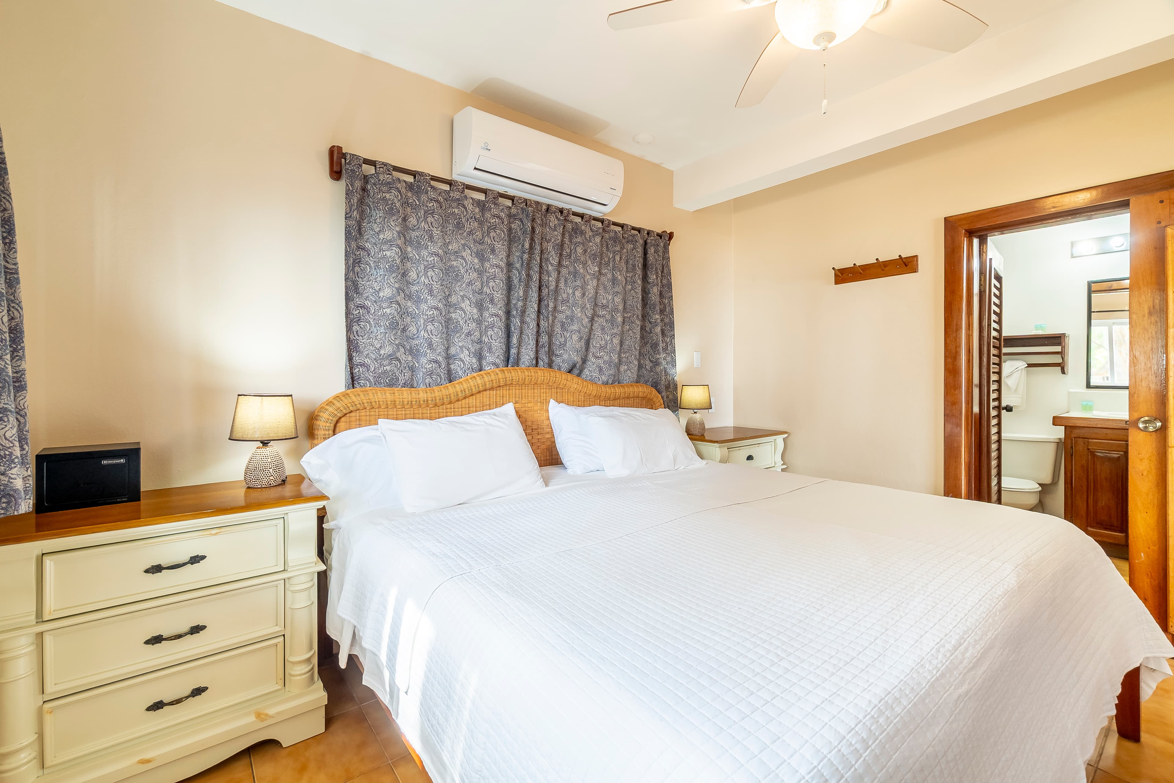 Modern bedroom in a tropical suite featuring a king-size bed with crisp white linens, a decorative wood headboard, and cream-colored bedside dressers. The room is equipped with a ceiling fan, air conditioning, and a view into an adjacent modern bathroom.