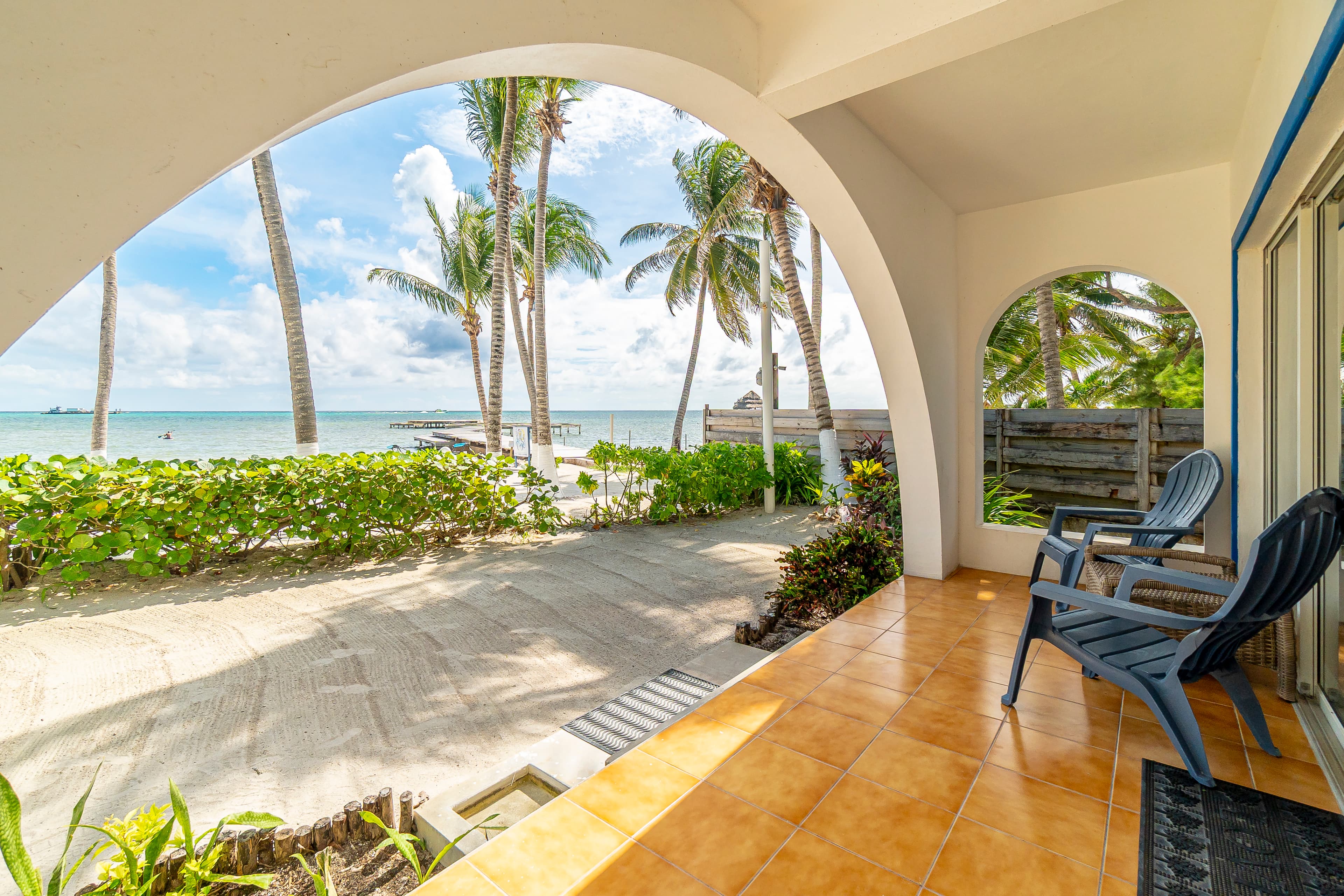 Ground-level private patio with tan floor tiles and blue lounge chairs, featuring white arched openings that look out onto a sandy beach area with tropical greenery, palm trees, and the ocean under a blue sky.