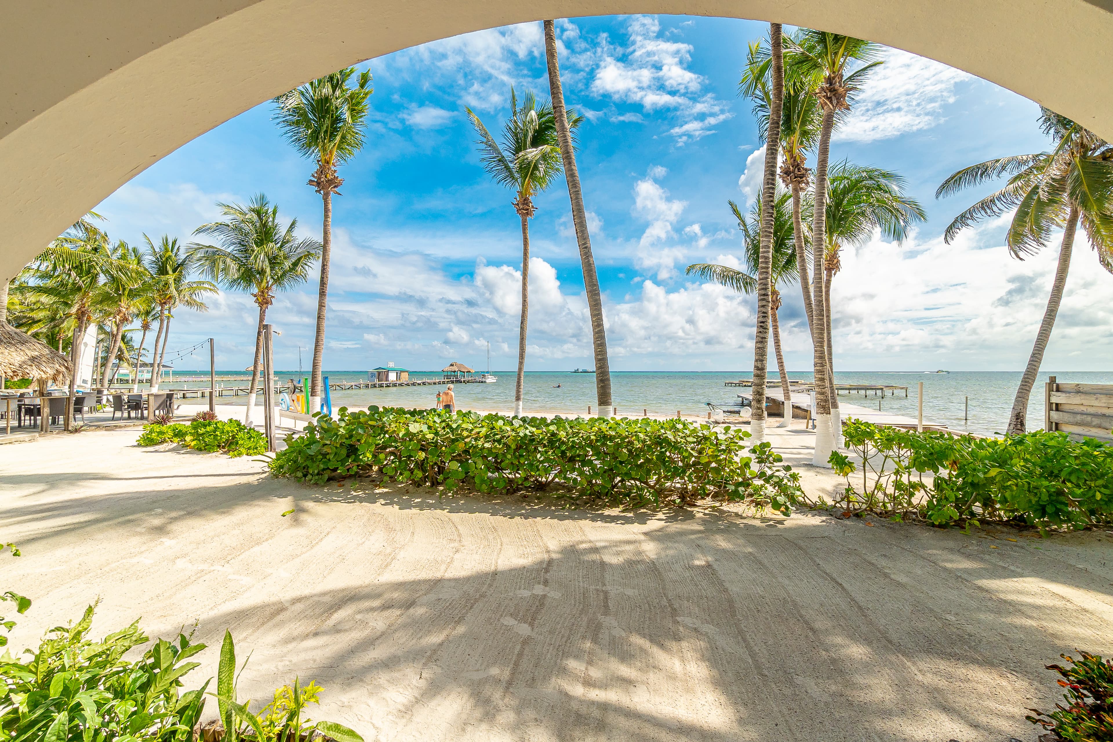 Beachfront view from a private patio with tan tiles and blue lounge chairs, looking through white arched openings to a sandy beach with palm trees and the blue Caribbean Sea.