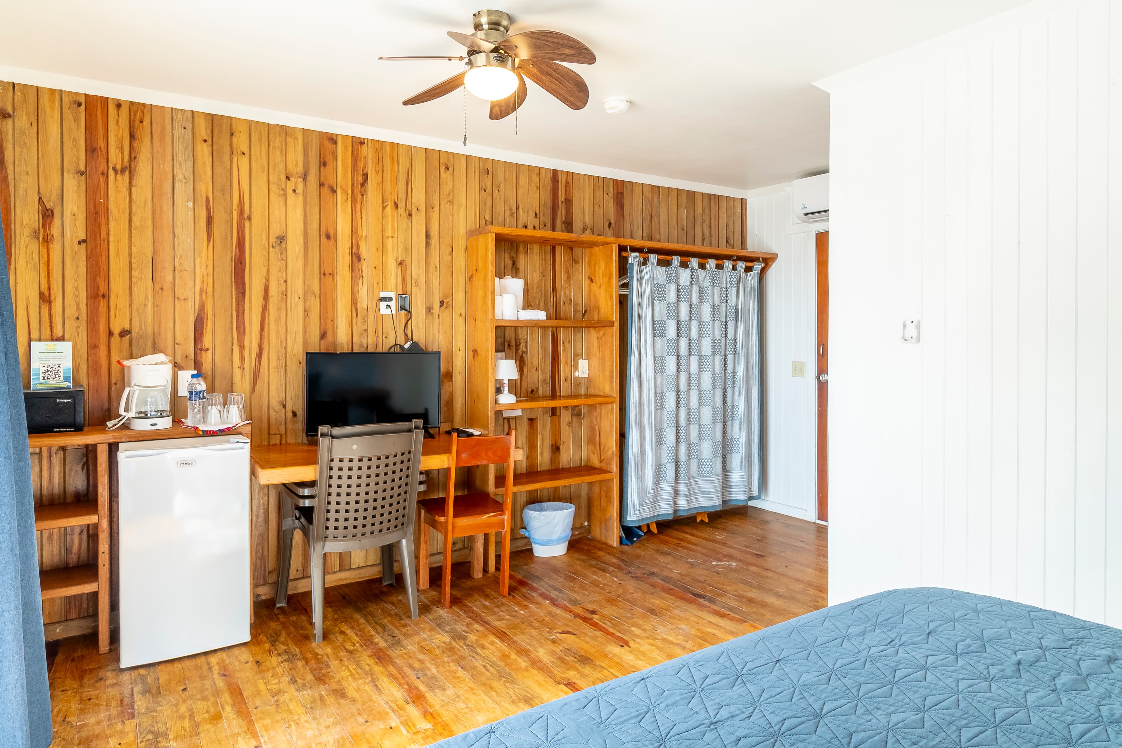Bedroom in a tropical suite featuring a king-size bed with a navy blue quilt and light blue wood-paneled walls. The room includes natural wood floors, a ceiling fan.