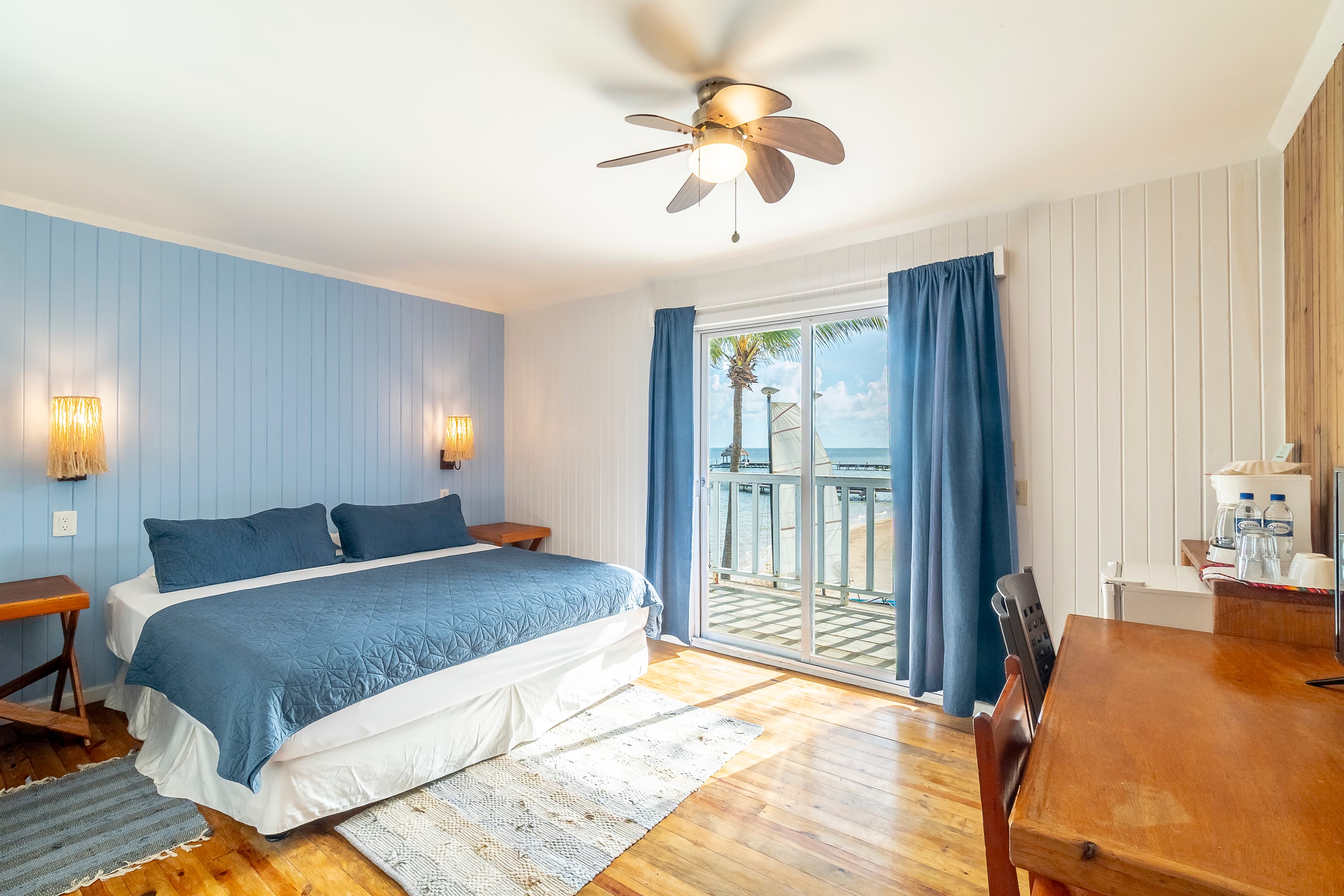 Modern beachfront bedroom featuring a king-size bed with a navy blue quilt, light blue wood-paneled walls, and large glass sliding doors that open to a balcony with views of the Caribbean Sea and palm trees.