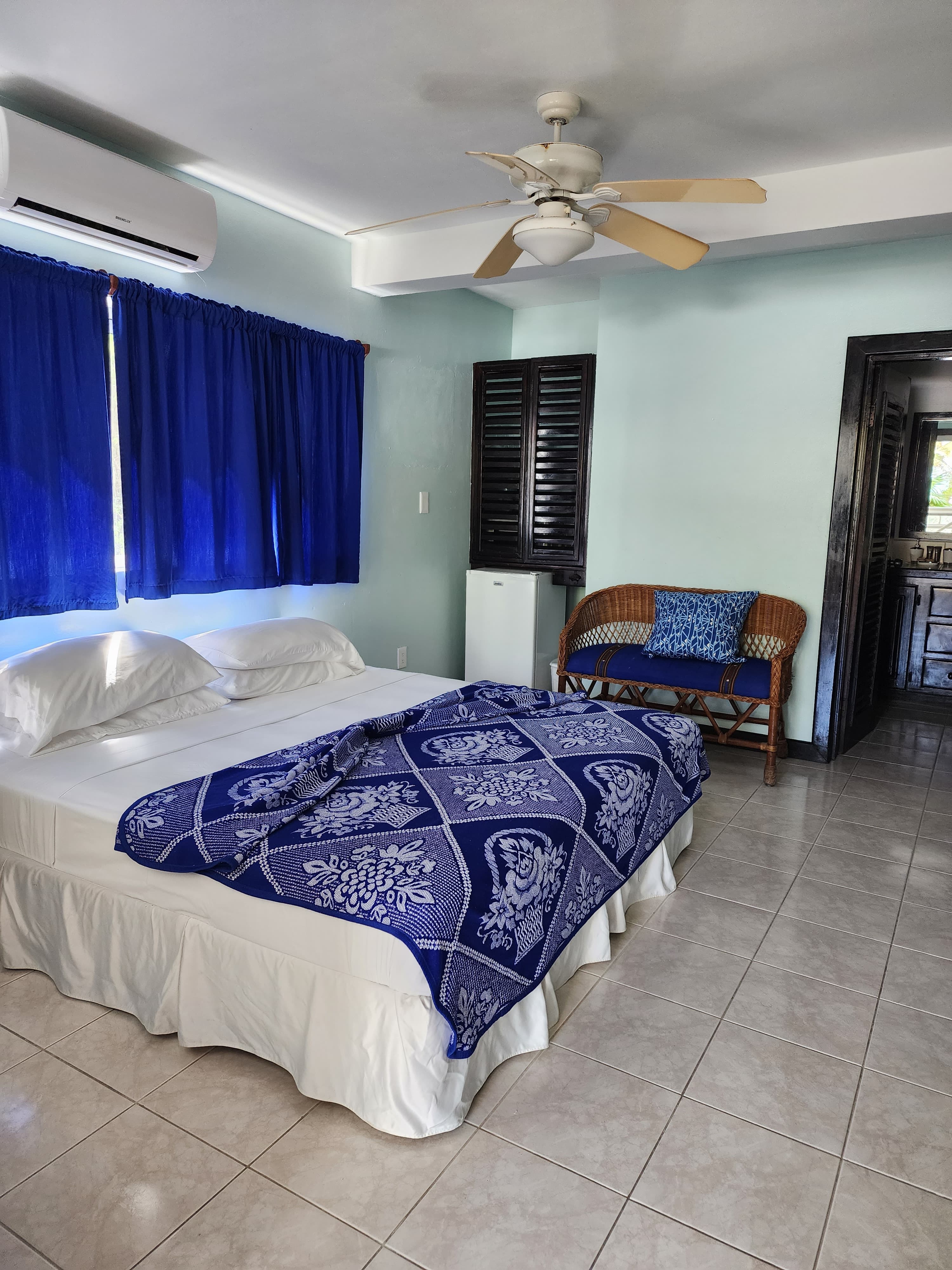 Bedroom in a tropical suite featuring a queen-size bed with a blue patterned quilt, light blue walls, a ceiling fan, and an air conditioning unit. The room includes tile flooring and a wicker bench with a matching blue cushion.