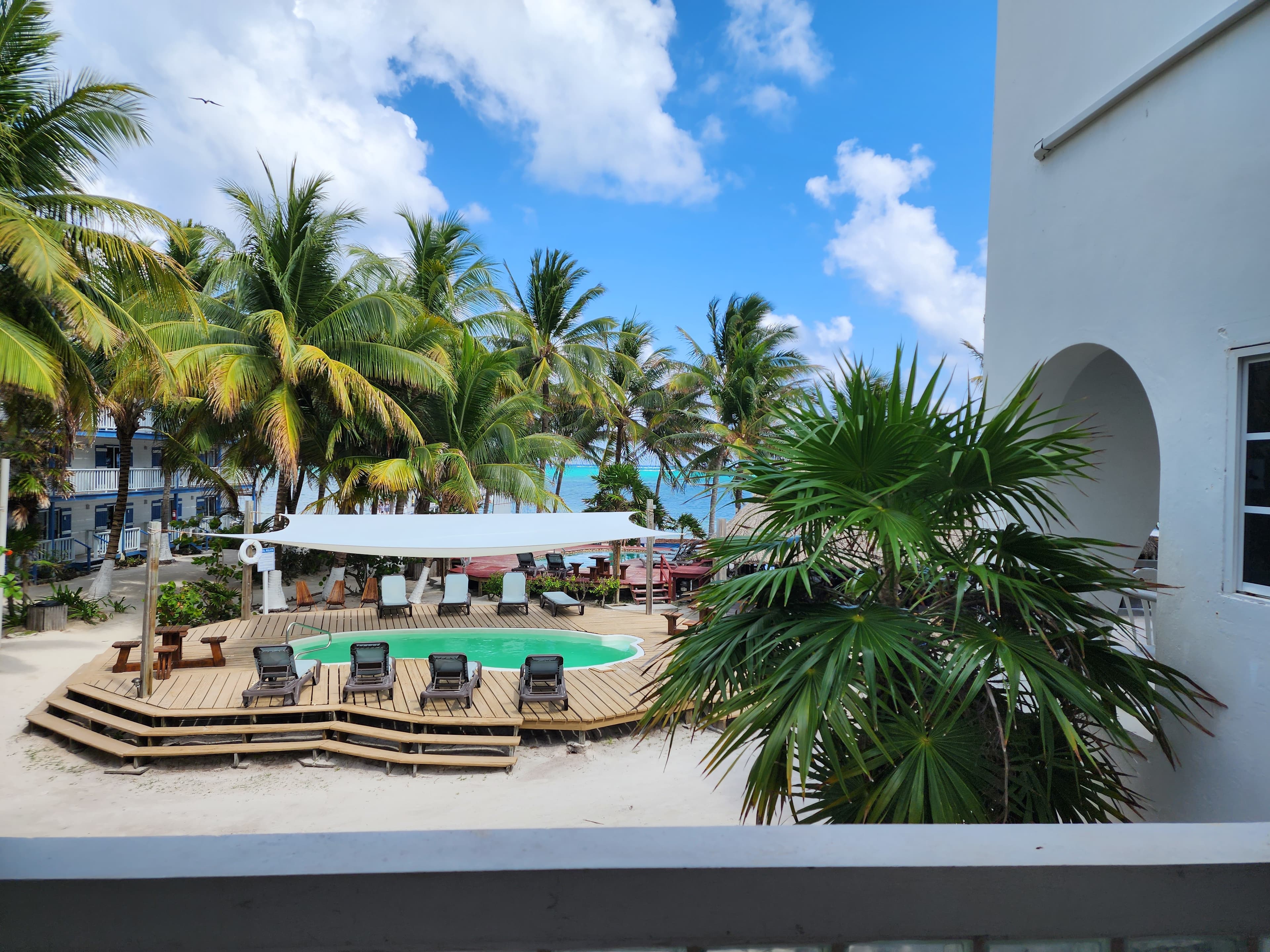 High-angle view of a tropical resort's outdoor swimming pool surrounded by a wooden deck with lounge chairs, palm trees, and white sand, with the Caribbean Sea visible in the distance under a blue, cloudy sky.