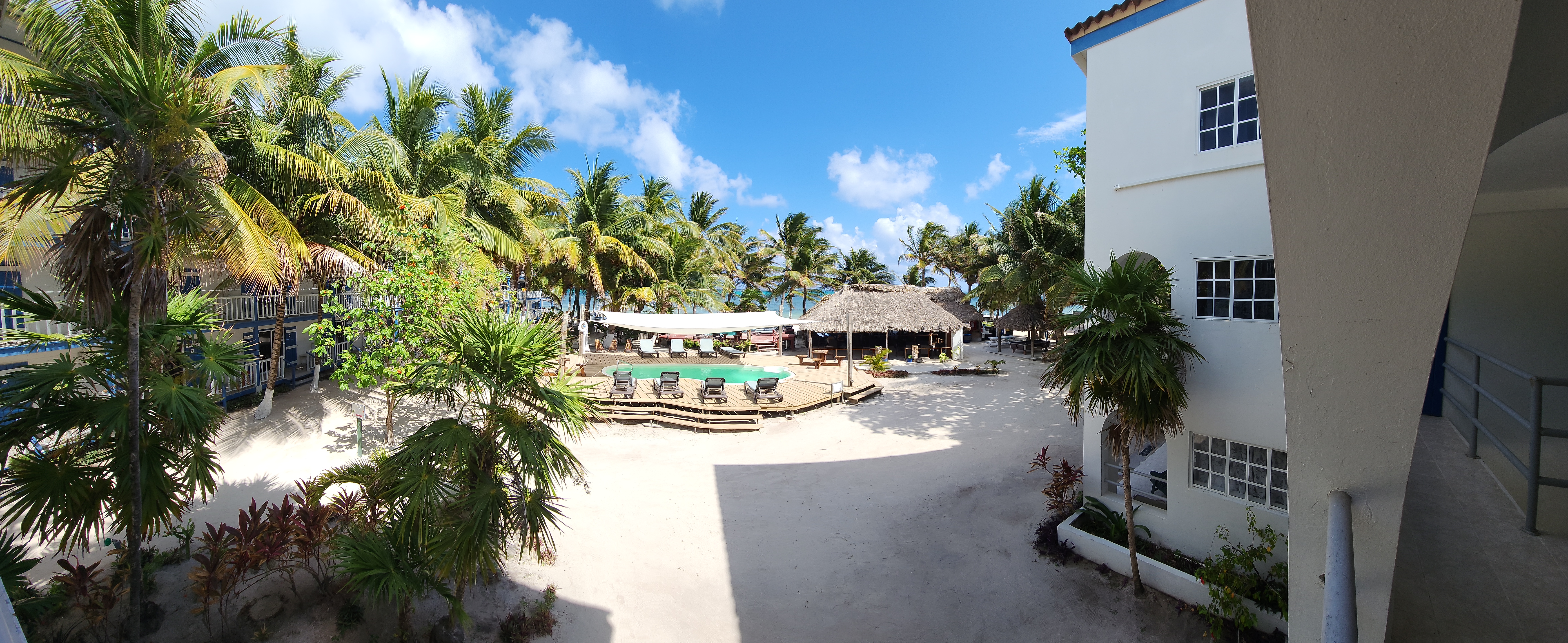 Wide-angle view of a tropical resort courtyard featuring a white-sand ground, a central swimming pool with a wooden deck and lounge chairs, and lush palm trees set against a blue sky with white clouds.