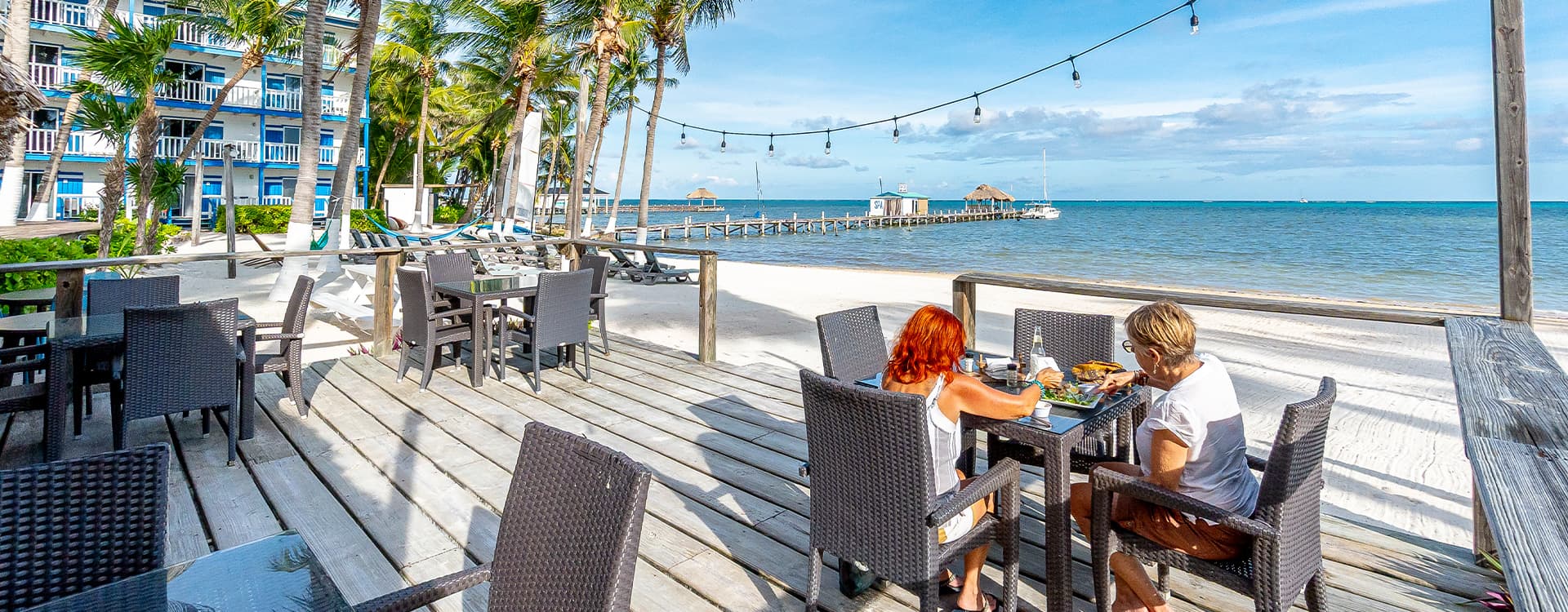 A couple enjoys a meal on a beachfront deck surrounded by palm trees and ocean views.