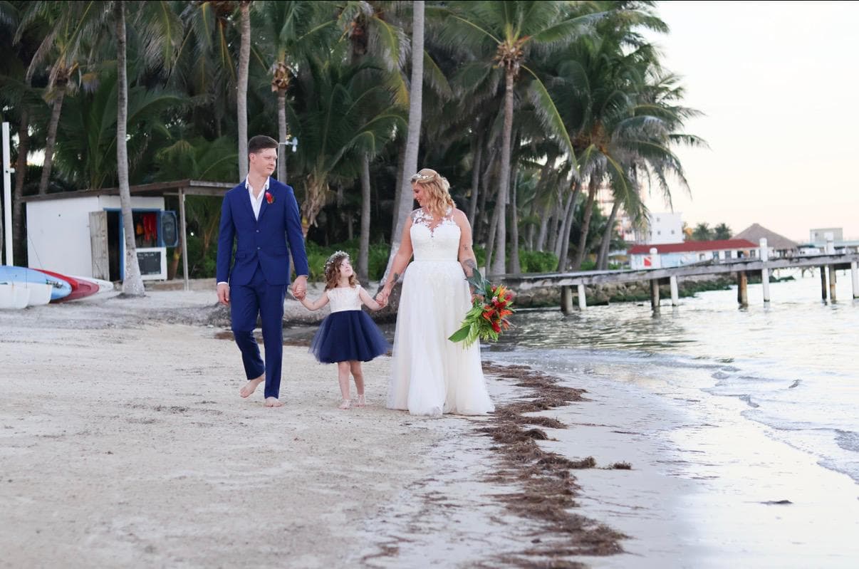 A couple in wedding attire walks hand-in-hand with a young girl along a beach lined with palm trees.