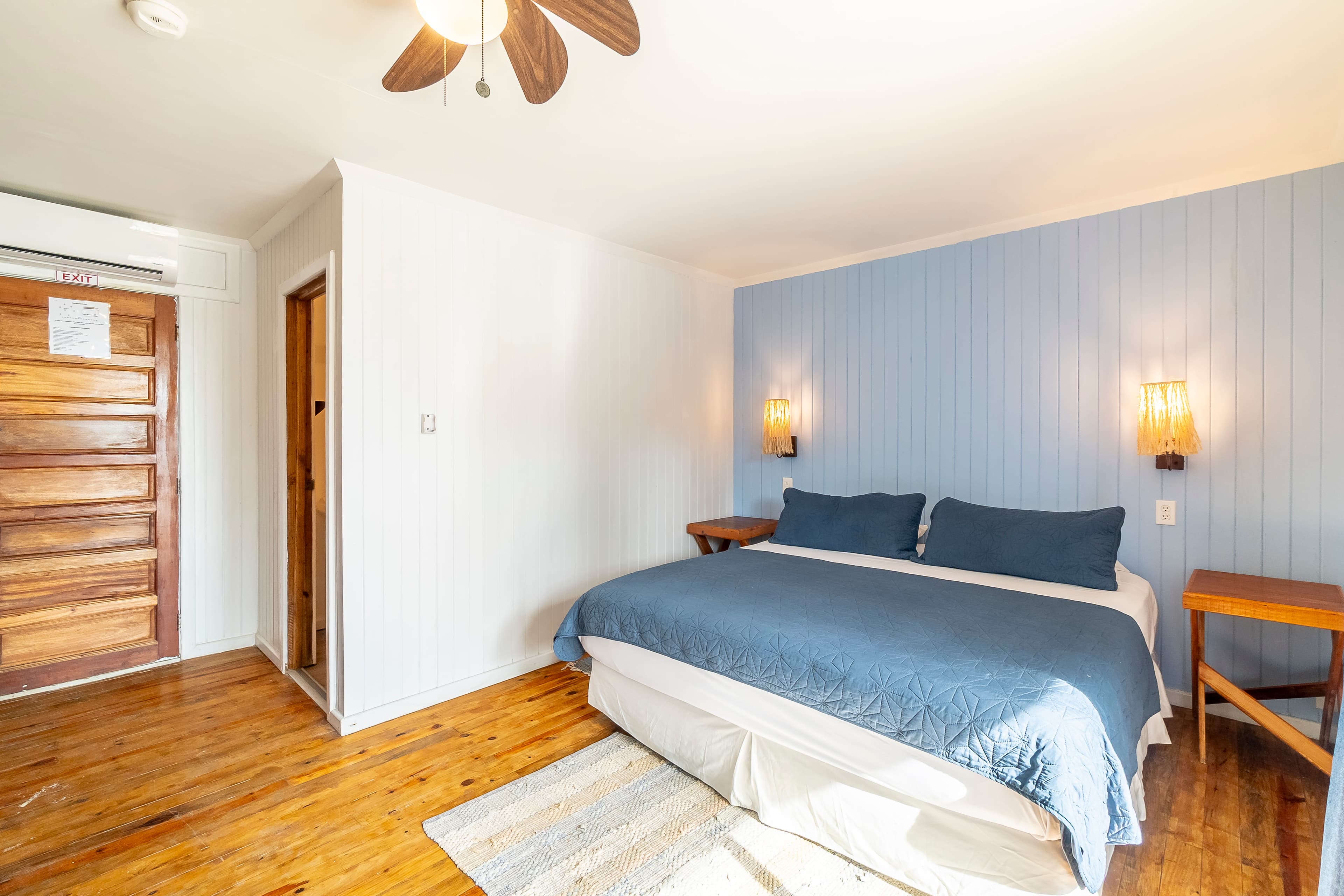 Modern beachfront bedroom featuring a king-size bed with a navy blue quilt, light blue wood-paneled walls, and large glass sliding doors that open to a balcony with views of the Caribbean Sea and palm trees.