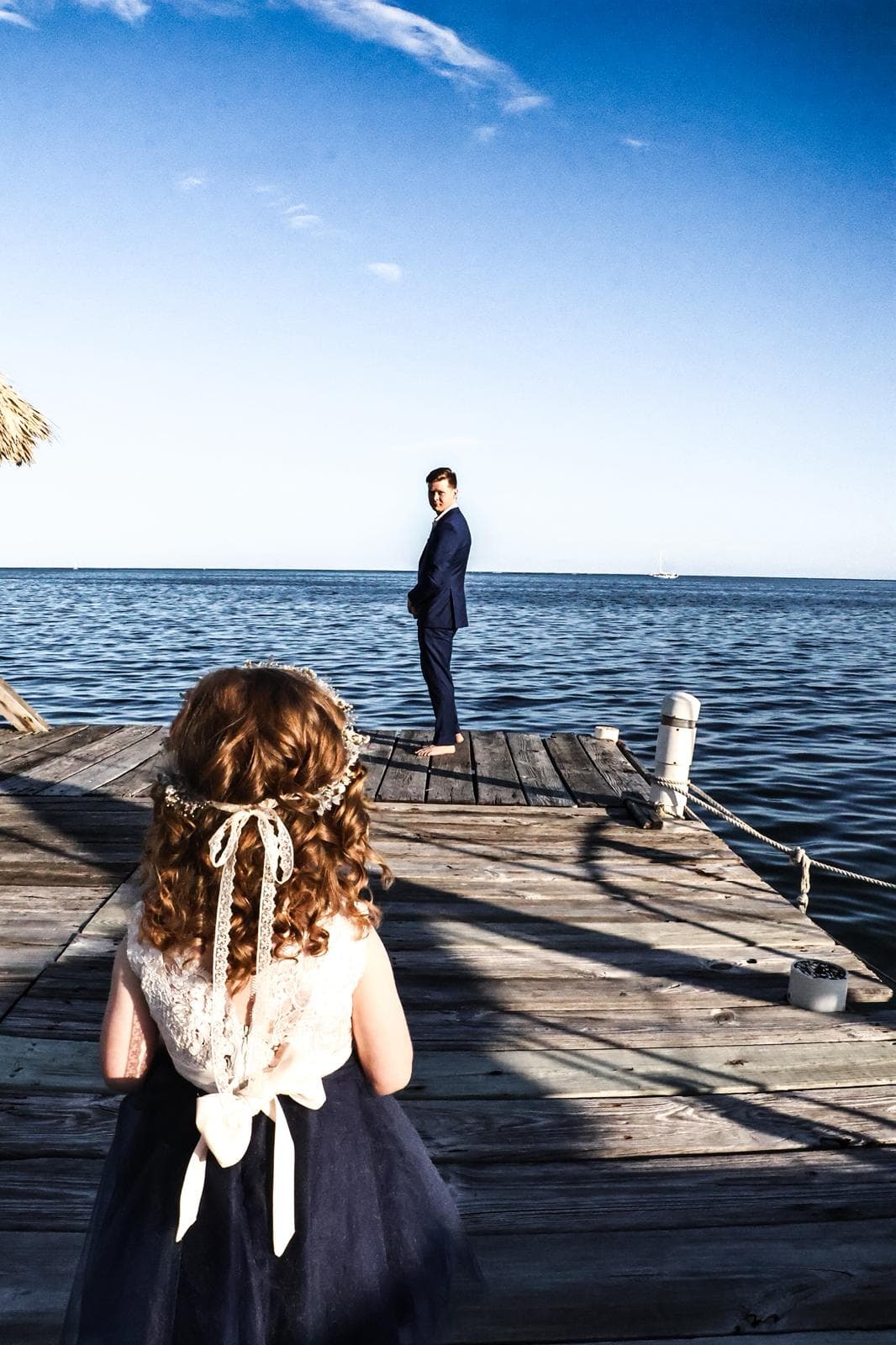 A young girl in a dress stands on a dock, looking at a man in a suit by the water.