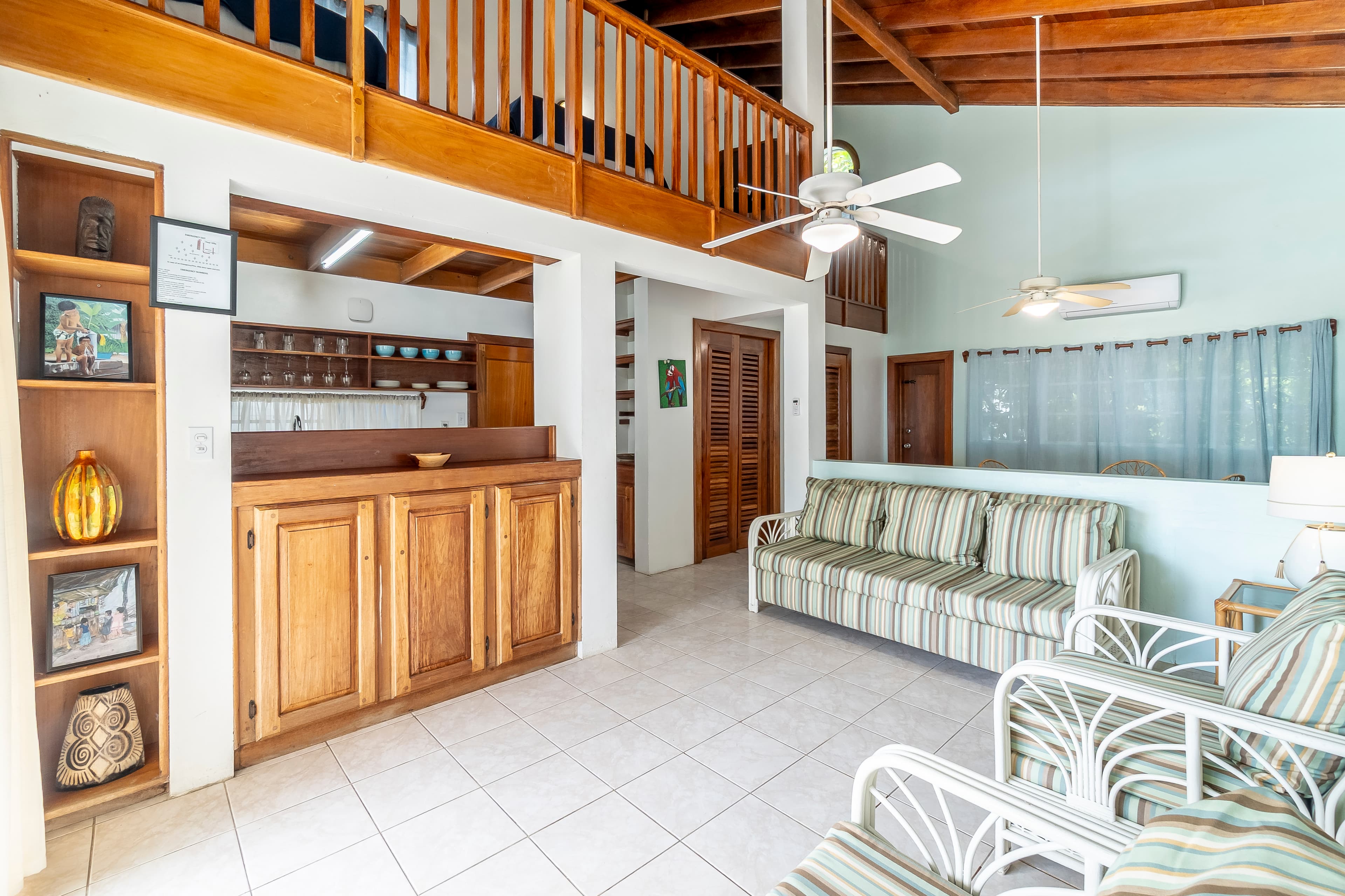 Spacious living area featuring high vaulted ceilings with wood beams, striped chairs & sofa, a breakfast bar, and a wooden balcony railing overlooking the room.
