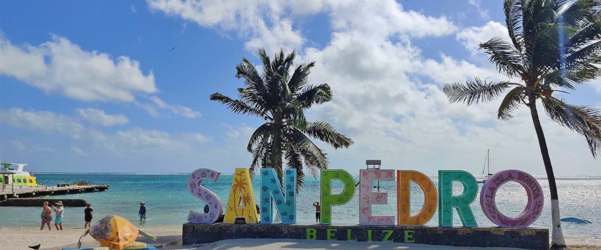 Colorful "San Pedro, Belize" sign in front of the beach with palm trees and a clear blue sky.