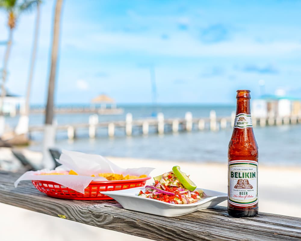 A bottle of Belikin beer beside a plate of seafood and a basket of nachos on a wooden railing overlooking the ocean.