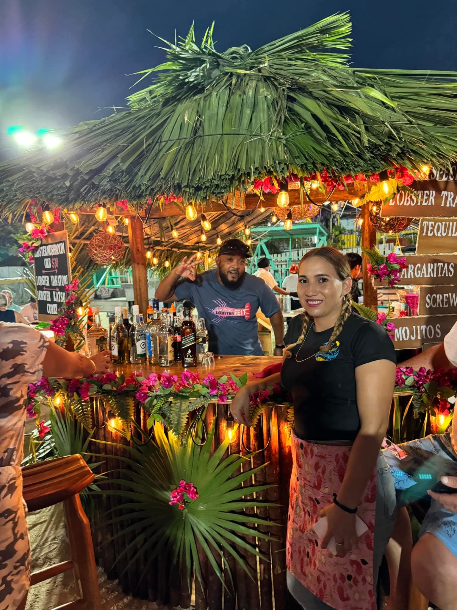 A vibrant outdoor bar scene at night with two staff members in front of a tiki-style bar adorned with tropical decor and various liquor bottles. A vibrant outdoor bar scene at night with two staff members in front of a tiki-style bar adorned with tropical decor and various liquor bottles.