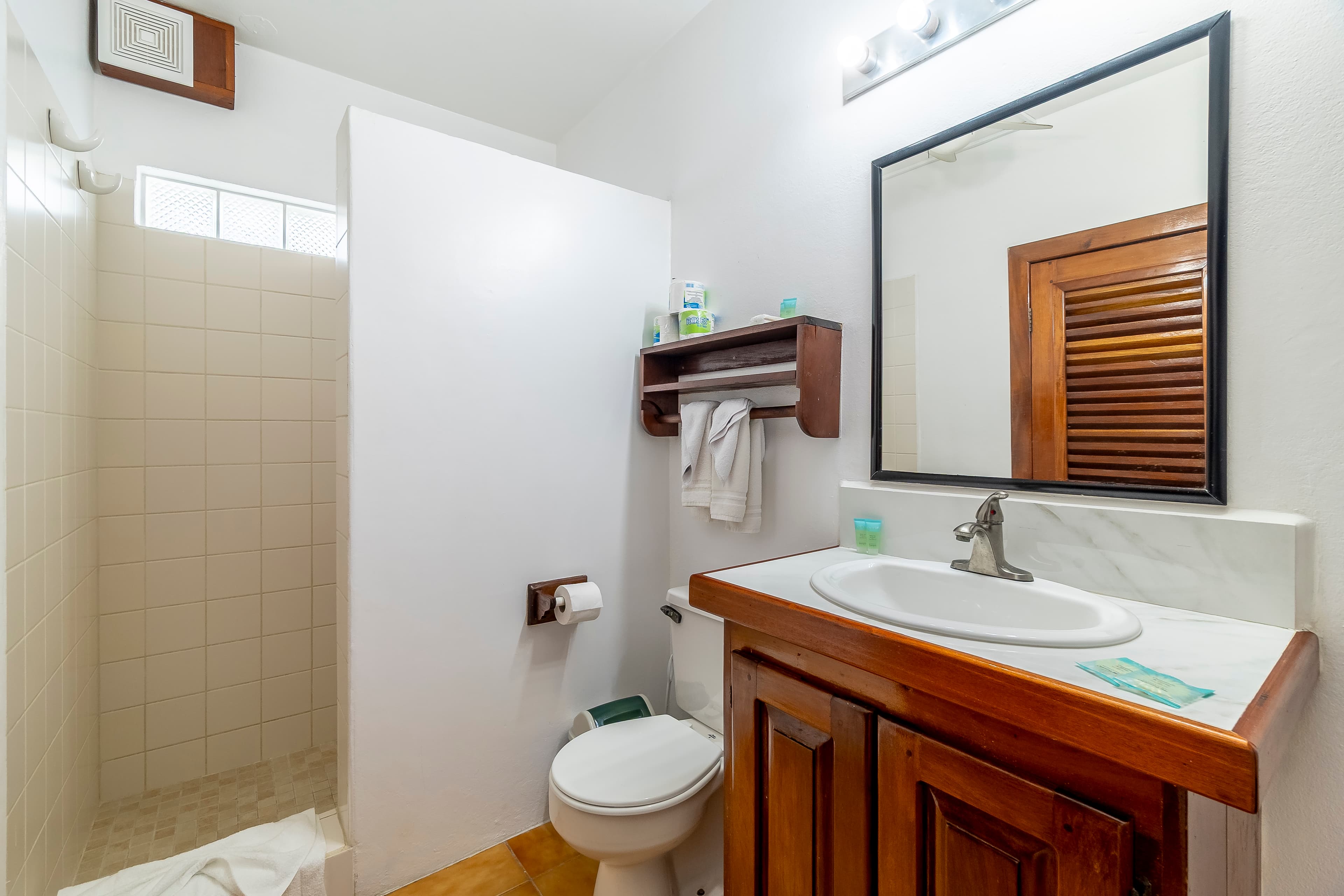 Clean, white-tiled bathroom in a tropical suite featuring a wooden vanity with a white countertop and sink, a large mirror with a black frame, a toilet, and a walk-in shower area.