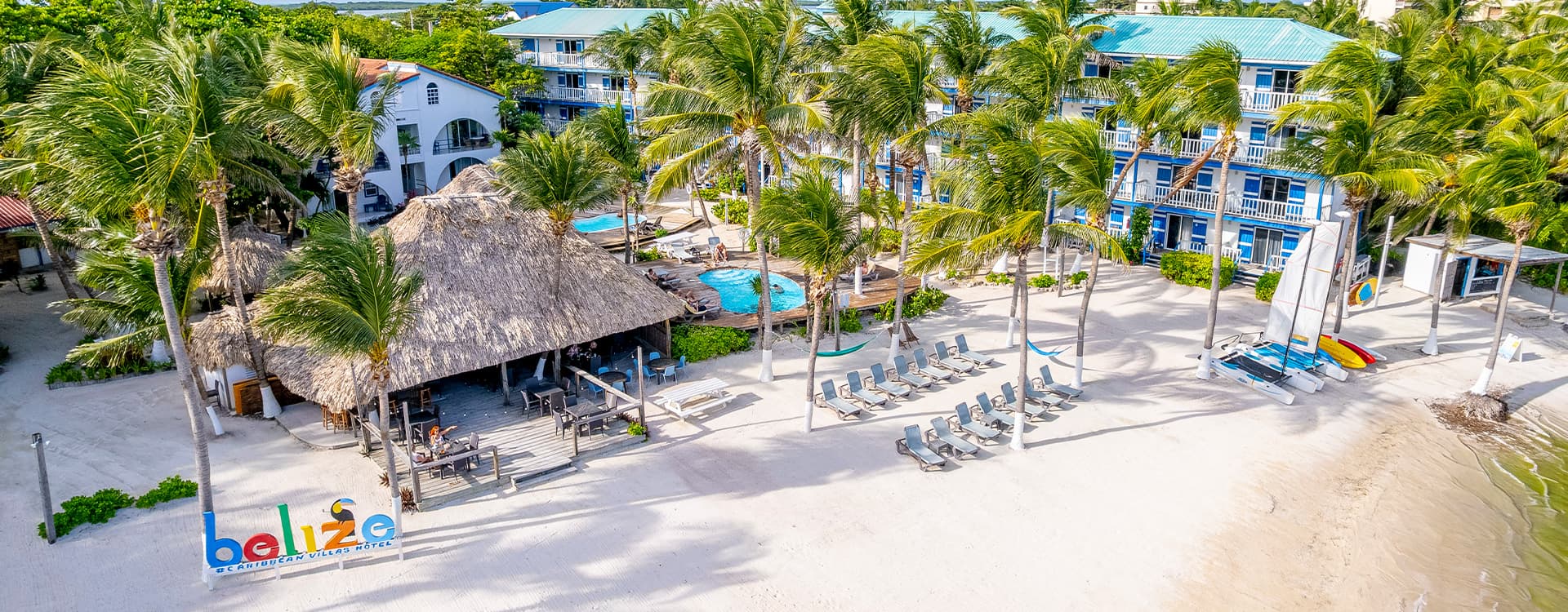 Aerial view of a beach resort in Belize featuring palm trees, a thatched-roof bar, swimming pools, and lounge chairs.