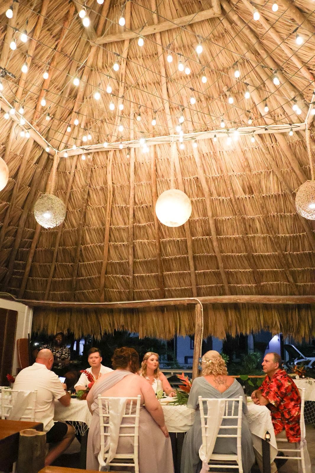 A group of people dining under a thatched roof illuminated by string lights.